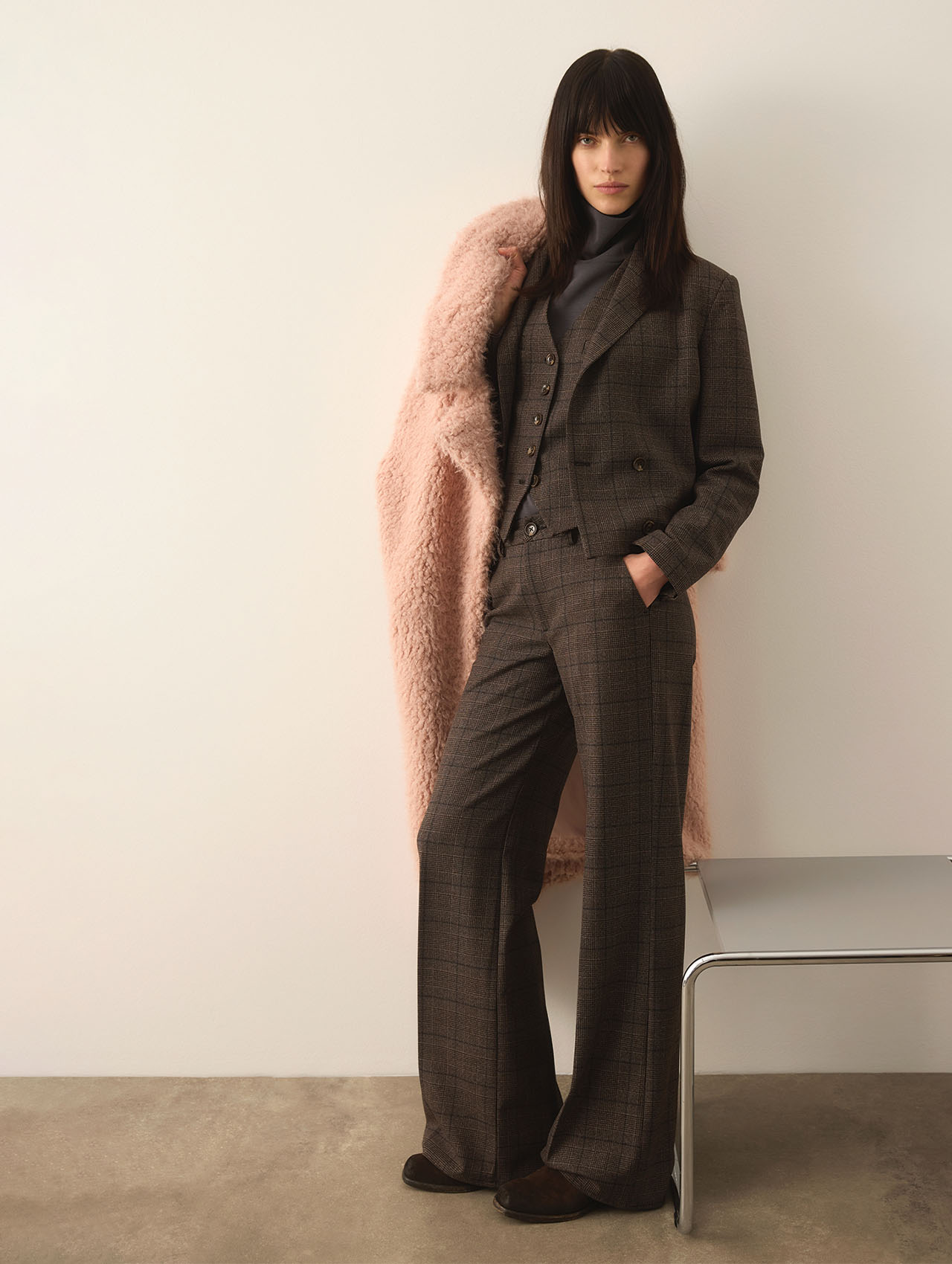 Eine Frau mit langen dunklen Haaren steht vor einer weißen Wand und trägt einen braunen karierten Anzug mit Weste und weit geschnittener Hose. Sie hat einen rosa Kunstpelzmantel über eine Schulter drapiert und steht neben einer minimalistischen Bank.
