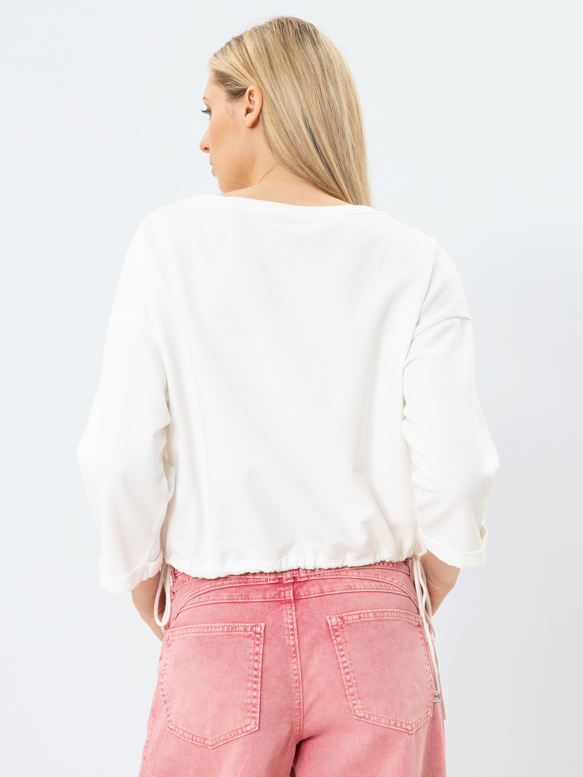 Eine Frau mit langen blonden Haaren ist von hinten zu sehen. Sie trägt ein locker sitzendes weißes Oberteil und hoch sitzende rosa Jeans. Sie steht vor einem schlichten, hellen Hintergrund.