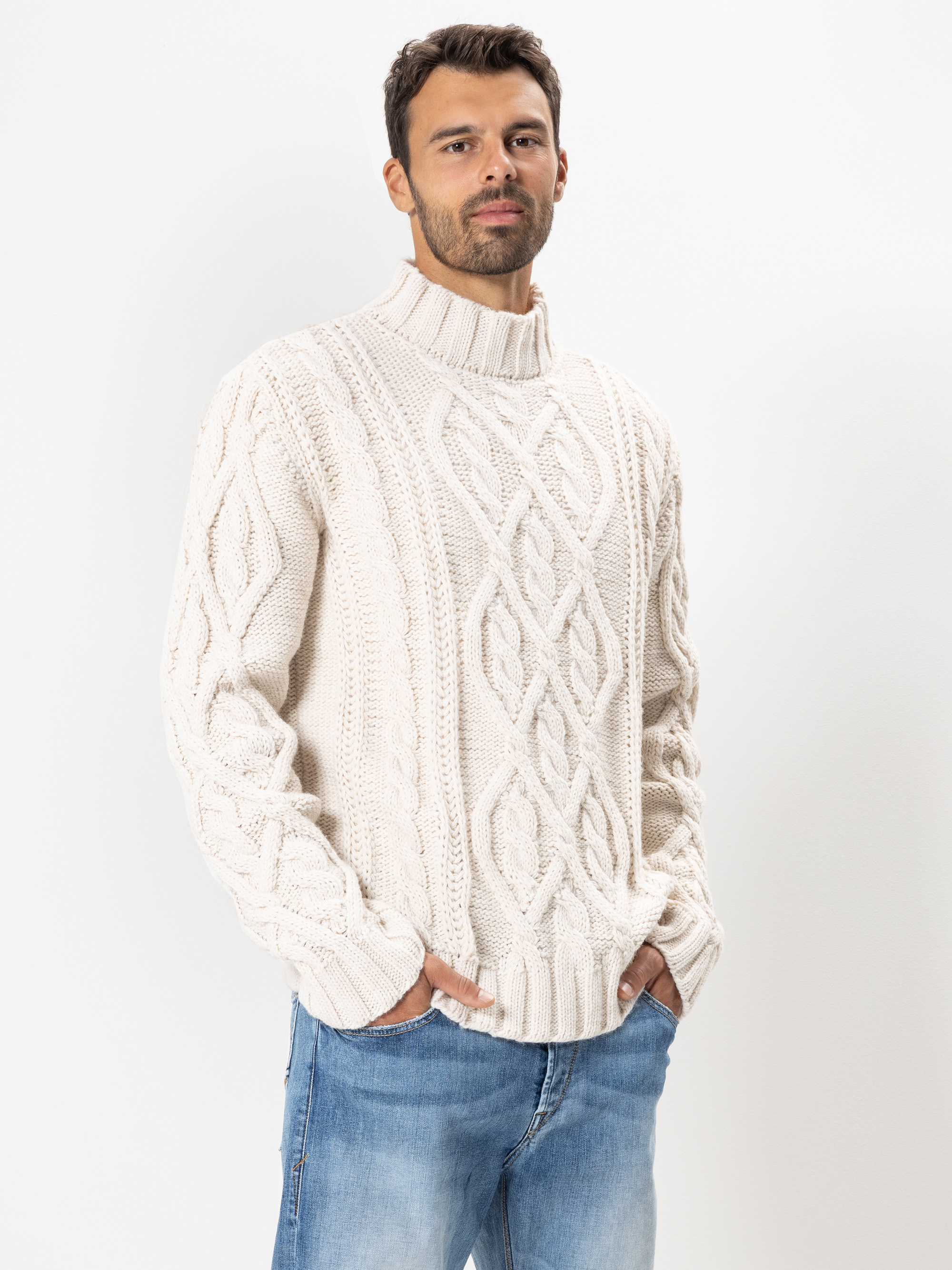 Ein Mann, der einen cremefarbenen, grob gestrickten Rollkragenpullover mit einem Rautenzopfmuster trägt, steht mit den Händen in den Taschen einer hellblauen Jeans vor einem schlichten weißen Hintergrund.