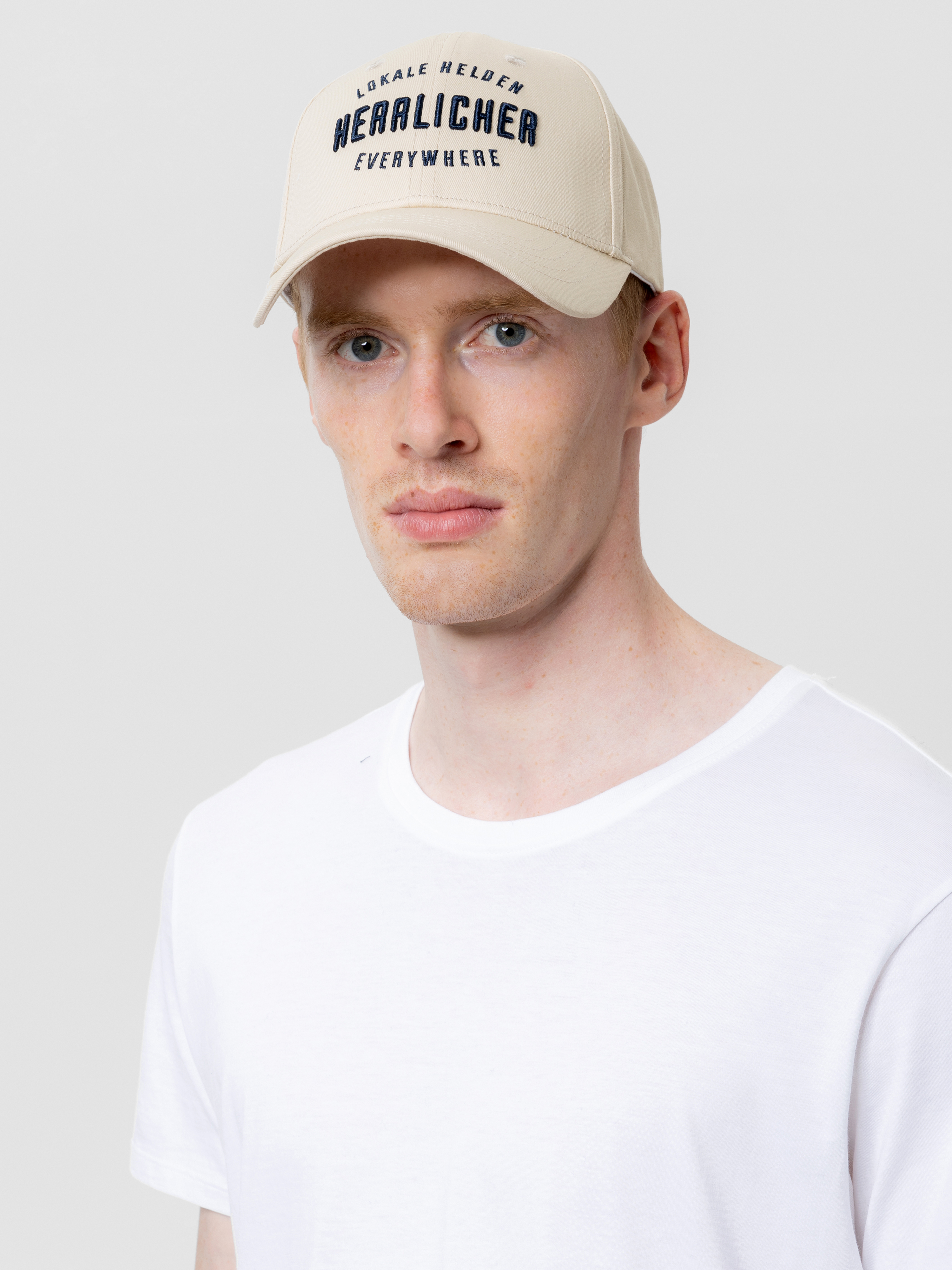 Ein Mann mit heller Haut und kurzen roten Haaren trägt eine beigefarbene Baseballkappe mit schwarzer Aufschrift und ein schlichtes weißes T-Shirt vor einem schlichten hellgrauen Hintergrund.