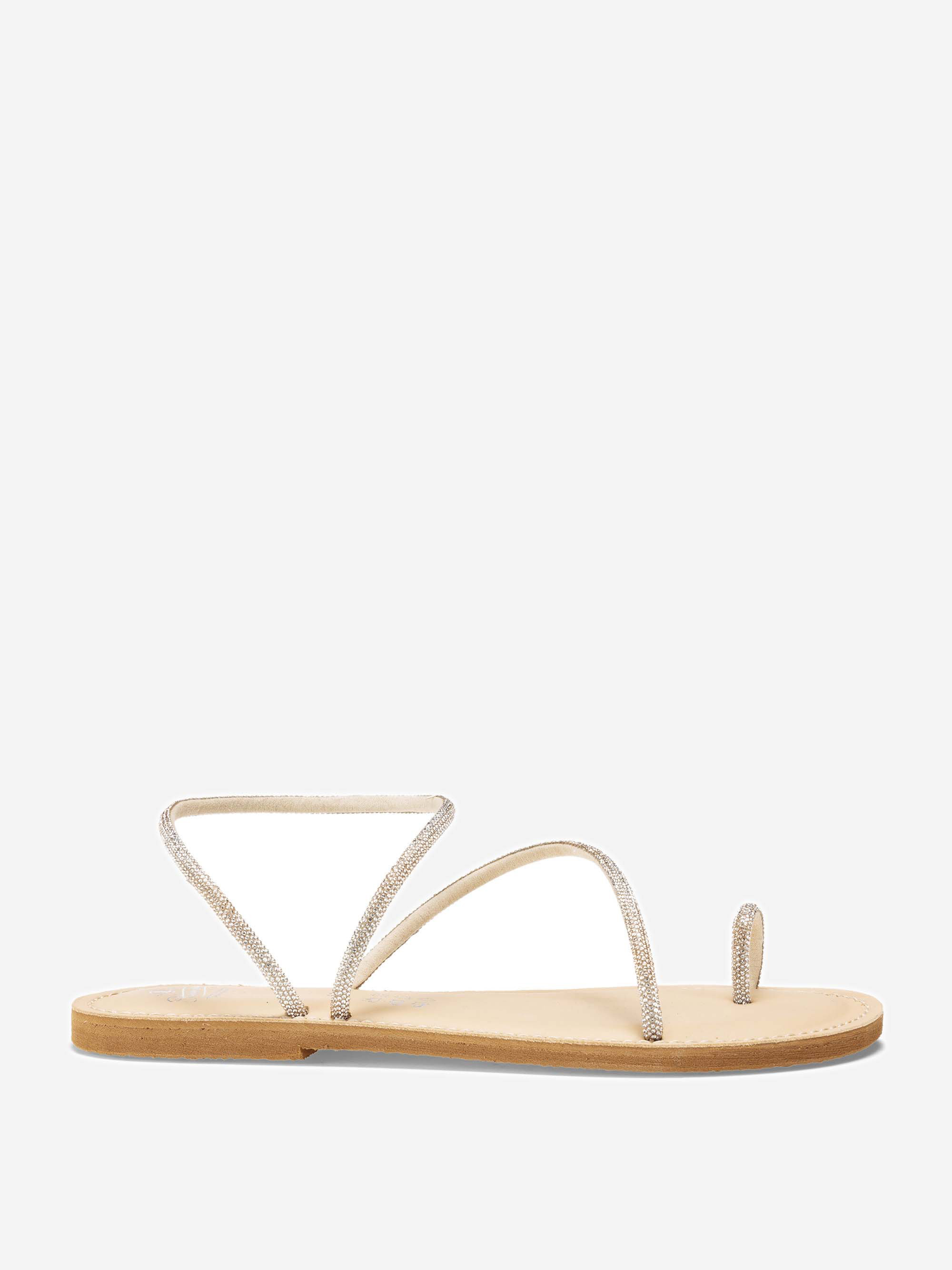 Eine minimalistische, flache Sandale mit dünnen, silbern glitzernden Riemen, einschließlich einer Zehenschlaufe und zwei diagonalen Riemen, auf einer hellbraunen Sohle vor einem schlichten weißen Hintergrund.