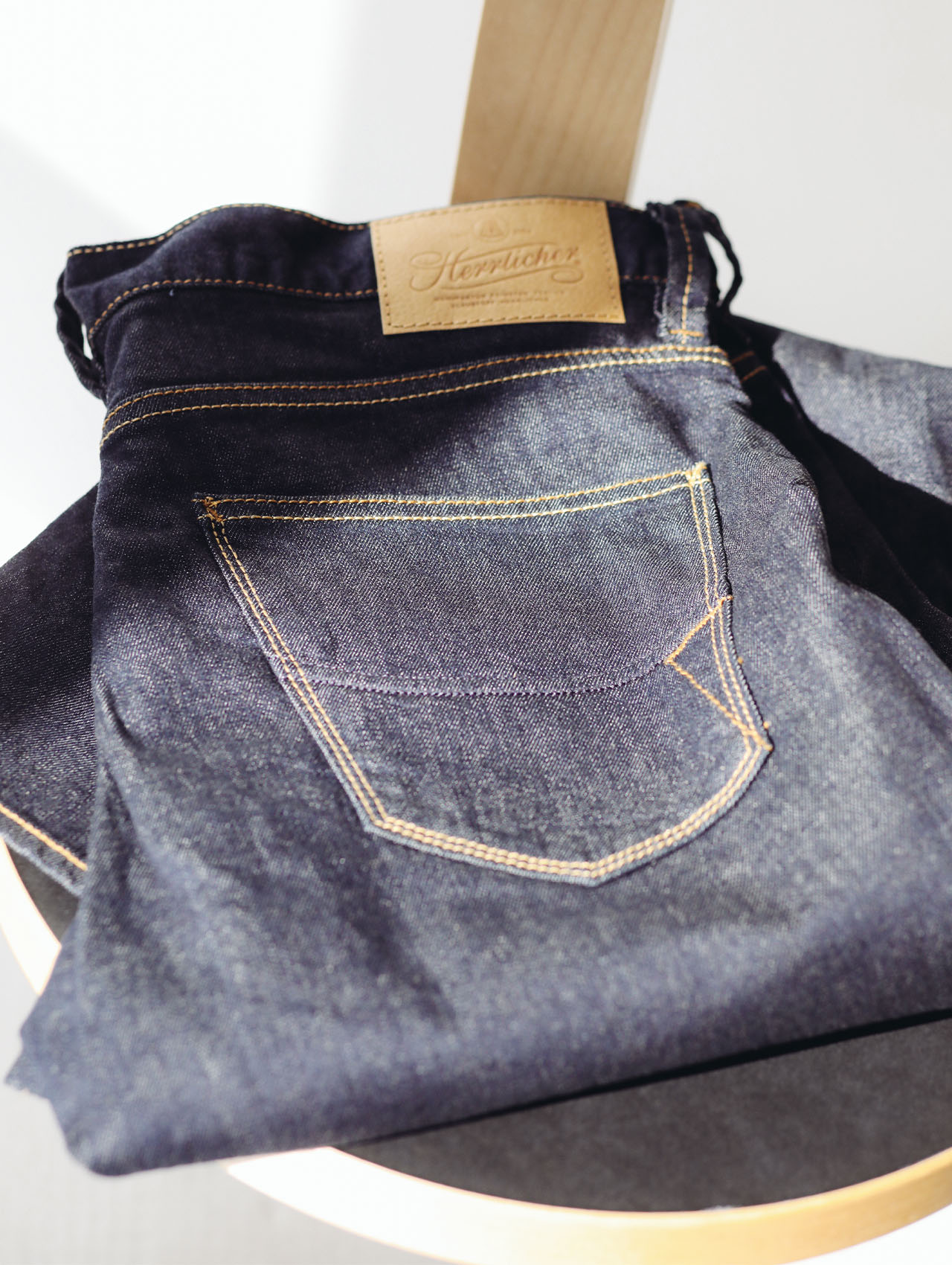 Eine dunkelblaue Jeans mit gelben Nähten und einem hellbraunen Lederaufnäher liegt ordentlich gefaltet auf einer runden Holzfläche im hellen Sonnenlicht.