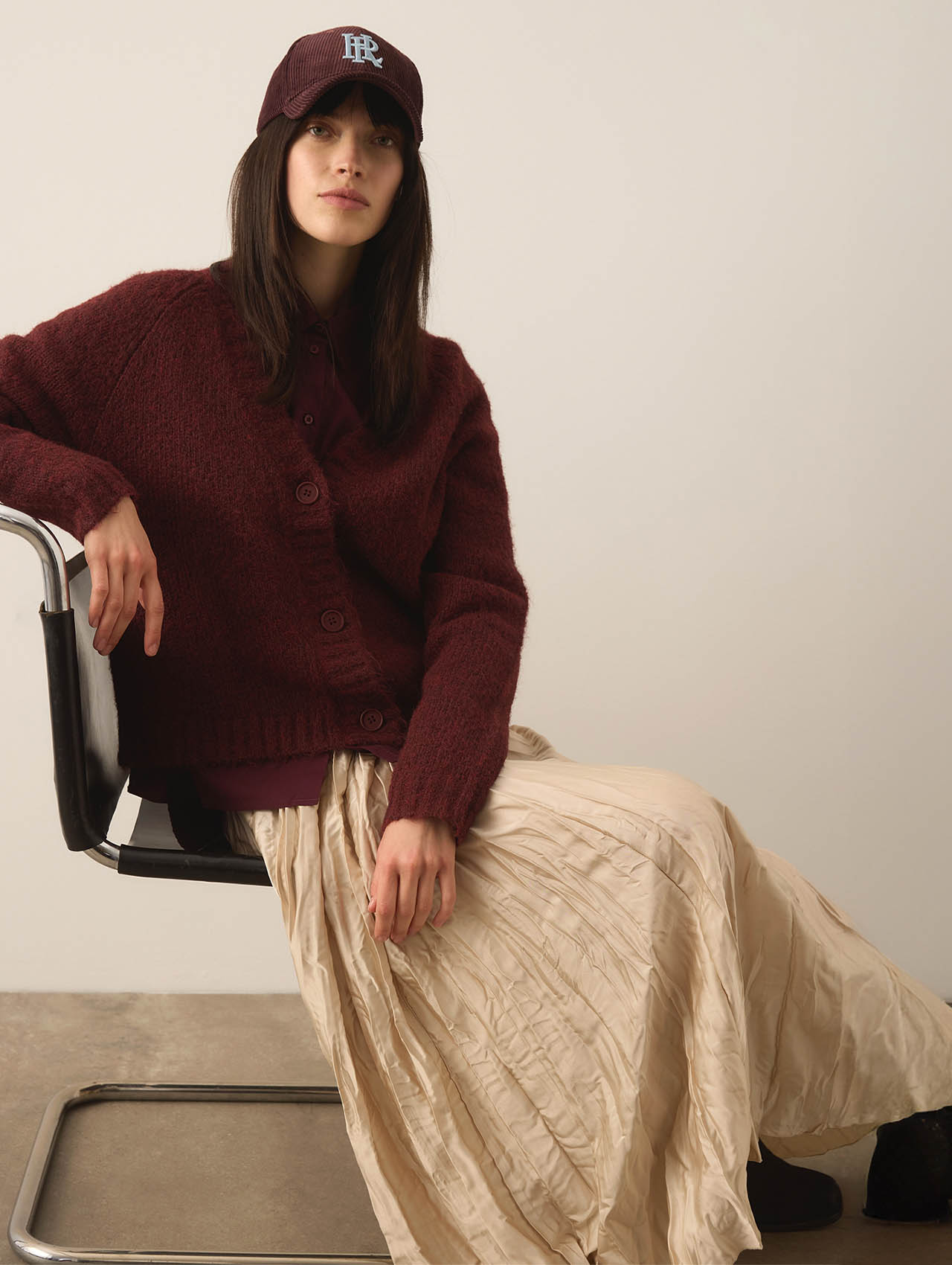 Eine Frau, die eine kastanienbraune Strickjacke, einen cremefarbenen Faltenrock und eine kastanienbraune Baseballmütze trägt, sitzt auf einem modernen schwarzen Stuhl vor einem schlichten Hintergrund.