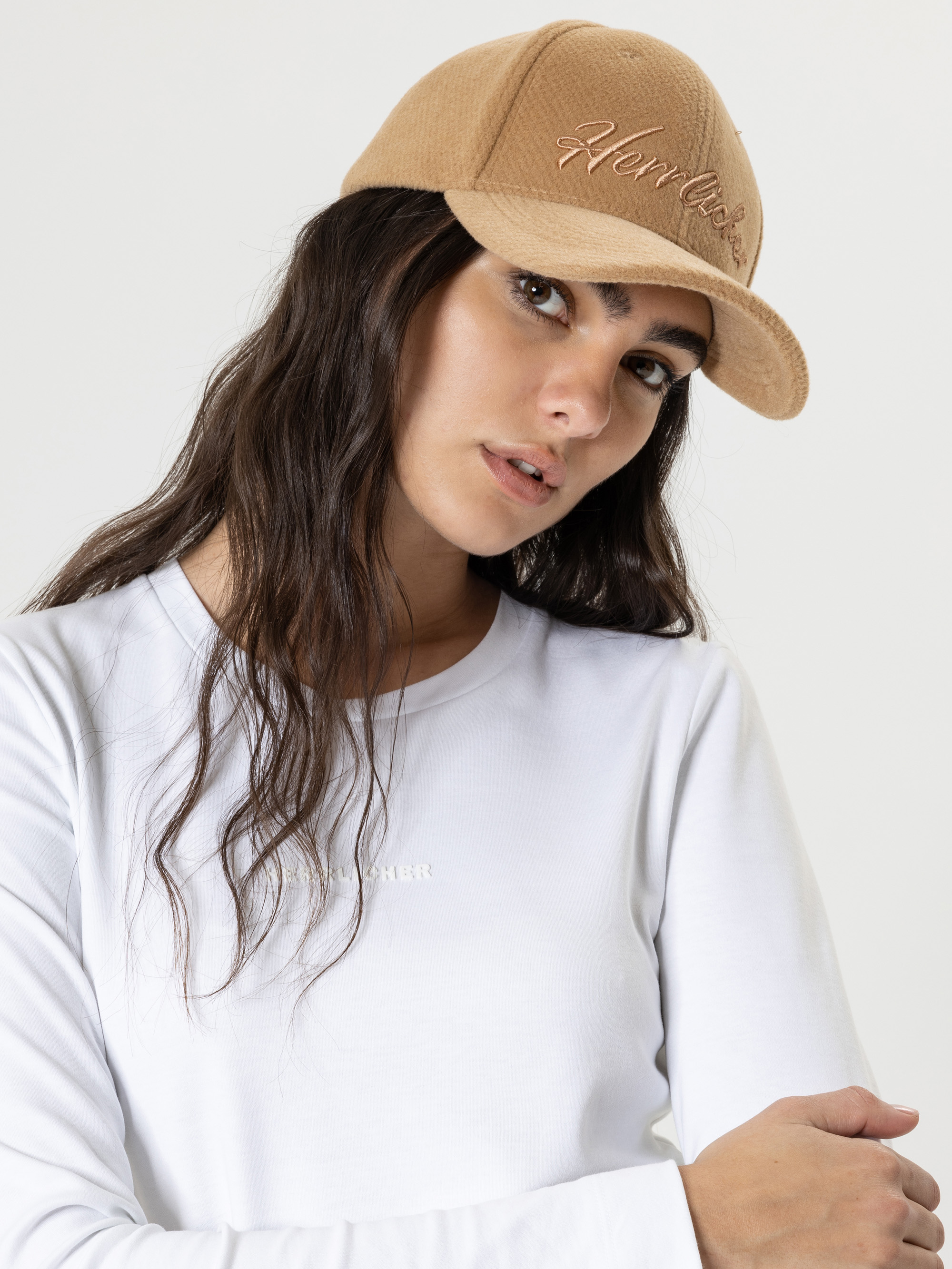 Eine Frau mit langen braunen Haaren trägt eine hellbraune Baseballkappe und ein weißes langärmeliges Hemd und posiert vor einem schlichten hellen Hintergrund. Sie blickt leicht zur Seite und hat einen neutralen Ausdruck.