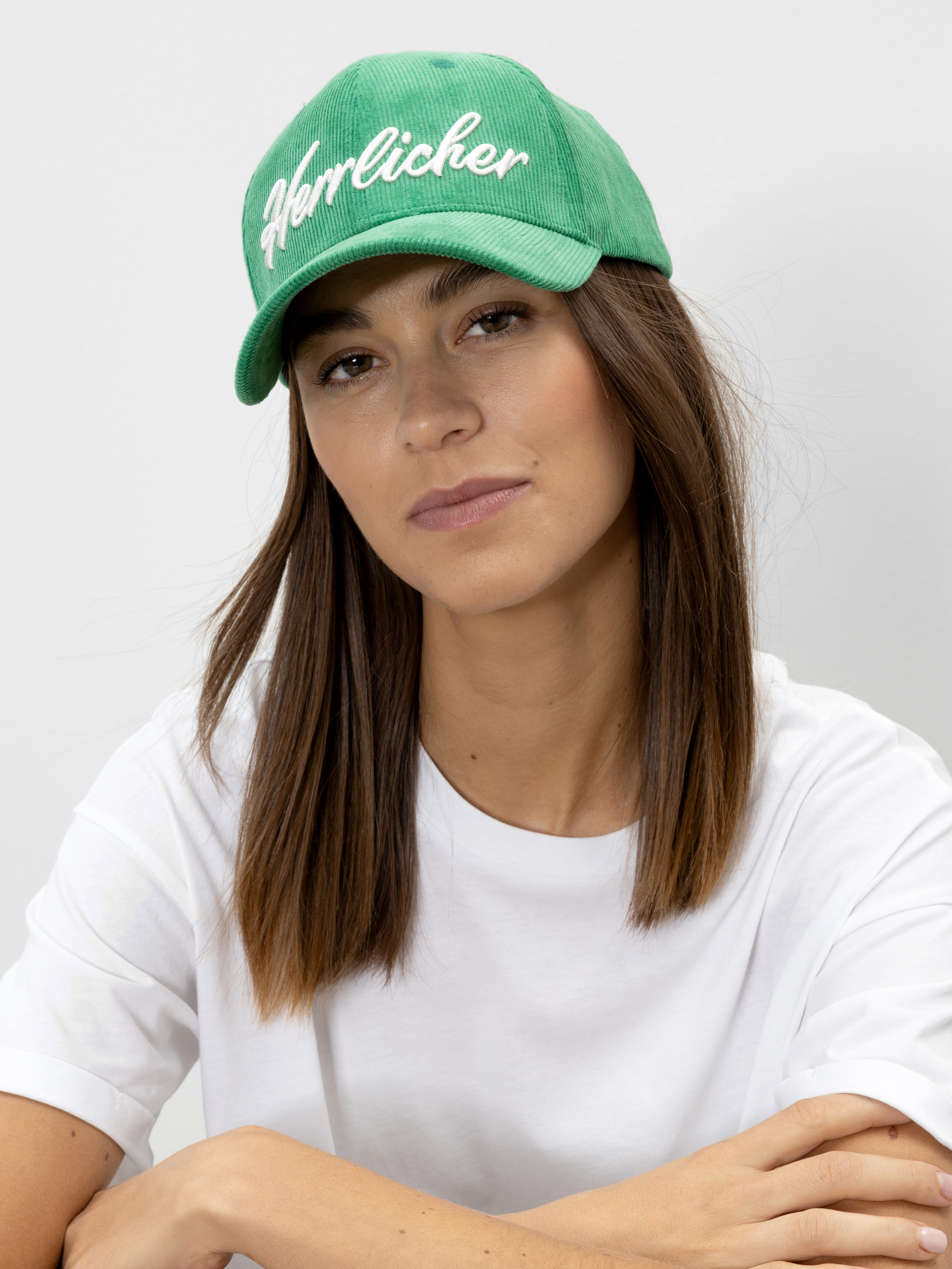 Eine Frau mit glattem braunem Haar trägt eine grüne Mütze mit weißem Herrlicher-Schriftzug und ein schlichtes weißes T-Shirt vor einem hellgrauen Hintergrund.