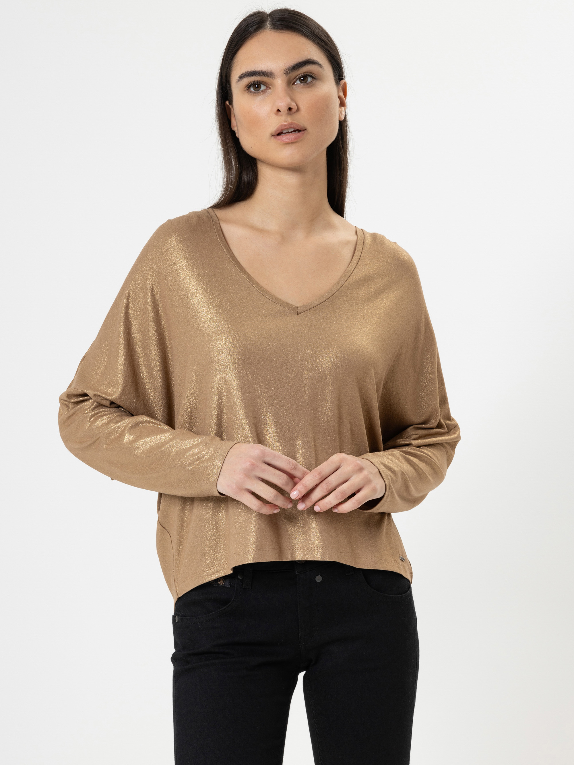 Eine Frau mit langen dunklen Haaren, die ein goldglänzendes langärmeliges Oberteil mit V-Ausschnitt und eine schwarze Hose trägt, steht vor einem schlichten weißen Hintergrund und blickt mit einem neutralen Ausdruck leicht nach oben.
