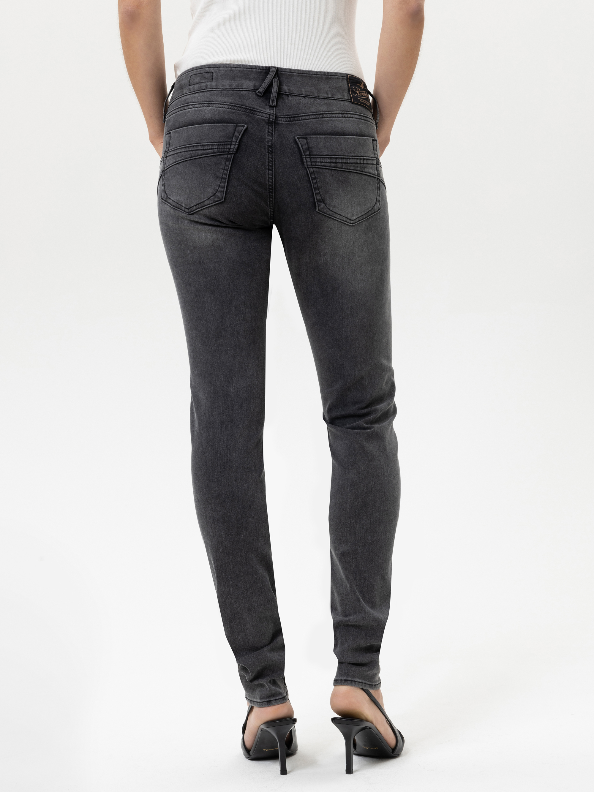 Person in dunkelgrauen Röhrenjeans und schwarzen hochhackigen Sandalen, stehend und von der Kamera abgewandt vor einem schlichten weißen Hintergrund.