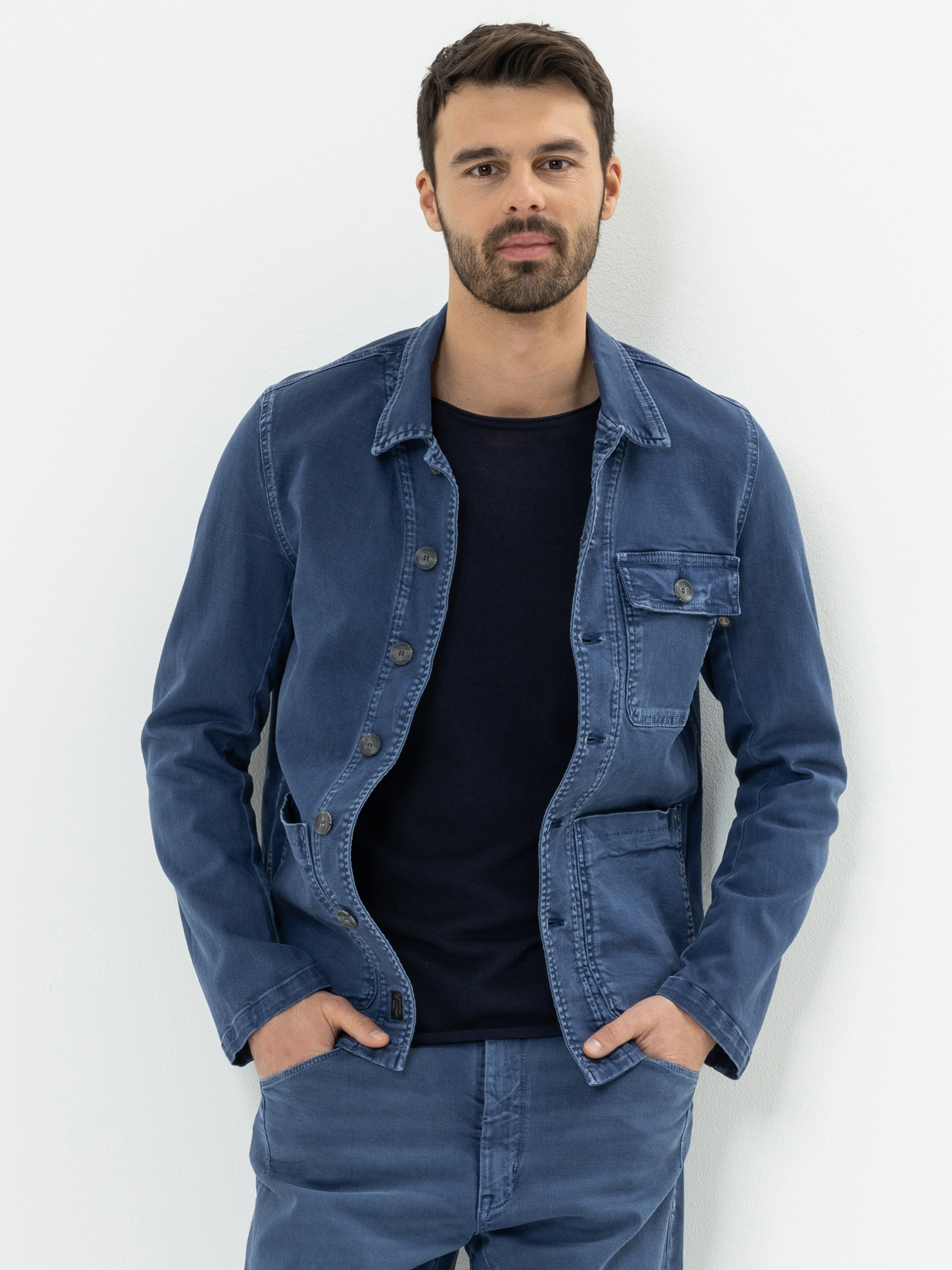 Ein Mann mit kurzen dunklen Haaren und einem Bart steht vor einer weißen Wand. Er trägt eine blaue Jeansjacke über einem schwarzen Hemd und dazu passende blaue Jeans, die Hände in den Taschen.