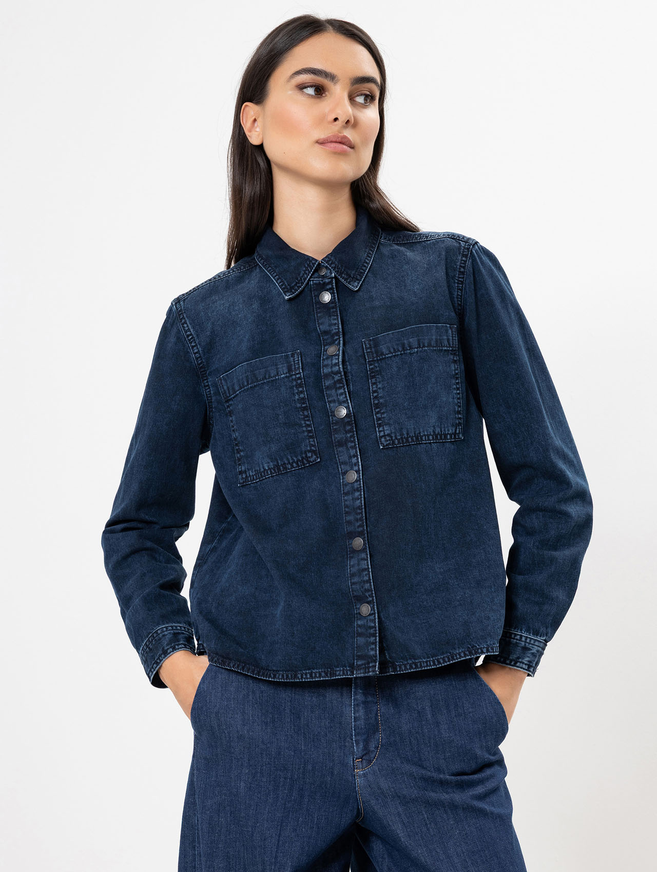 Eine Frau mit langen dunklen Haaren, die ein dunkelblaues Jeanshemd mit Knopfleiste und Vordertaschen und eine passende Jeanshose trägt, steht mit den Händen in den Taschen vor einem schlichten weißen Hintergrund.