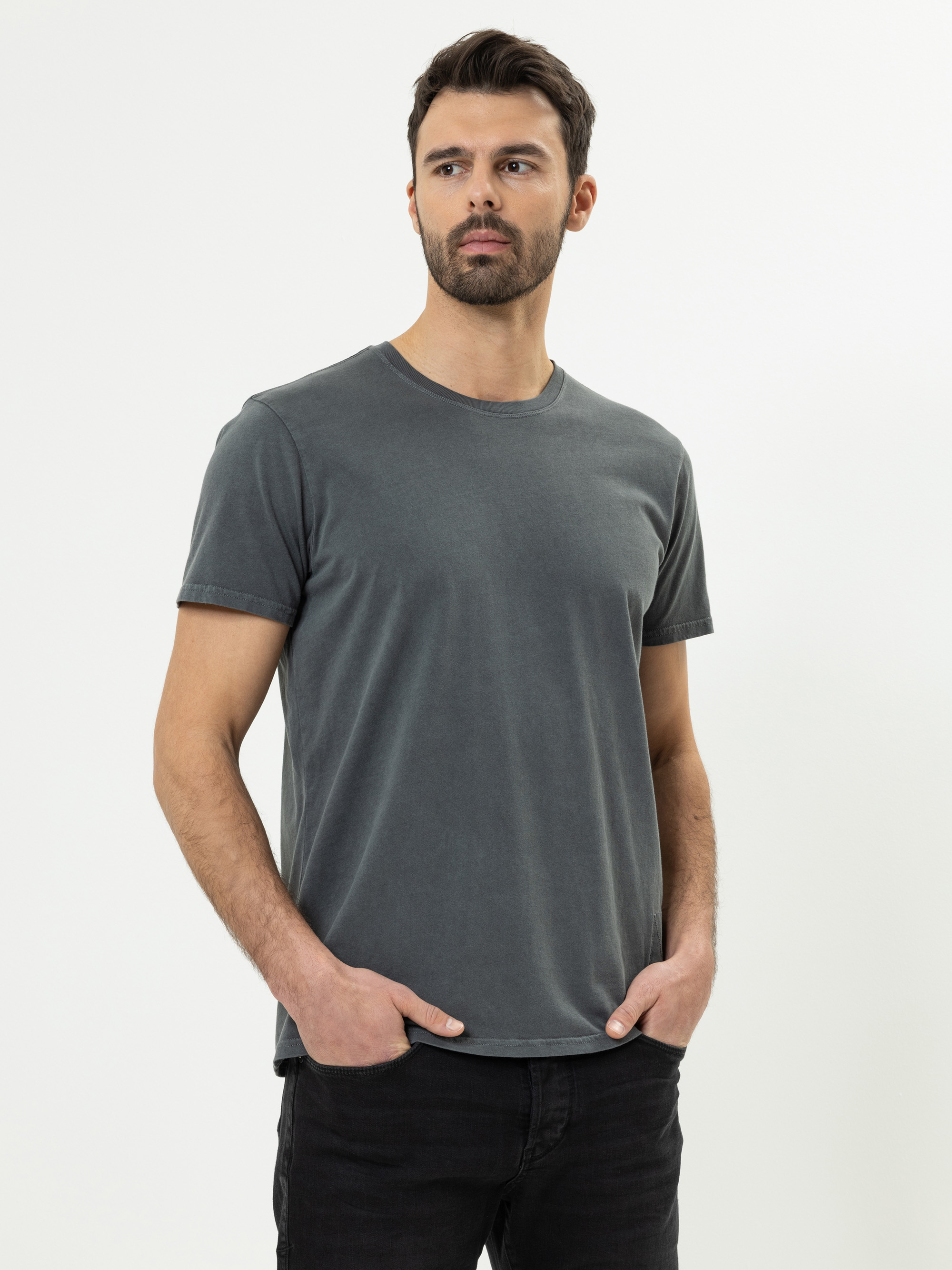 Vor einem schlichten weißen Hintergrund steht ein Mann mit kurzen dunklen Haaren und einem Bart, der ein schlichtes dunkelgraues T-Shirt und eine schwarze Hose trägt, die Hände in den Taschen hat und leicht zur Seite schaut.