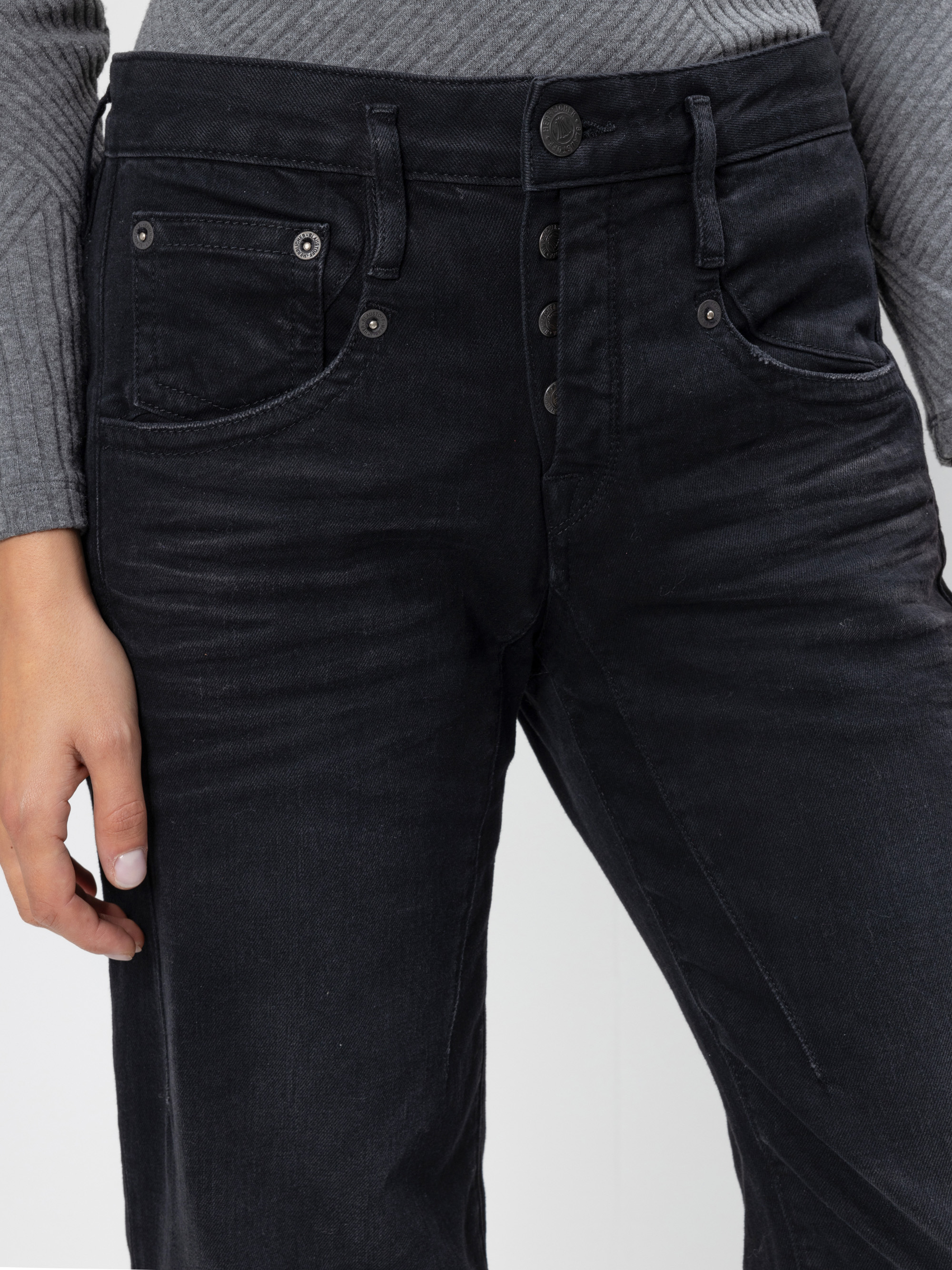 Eine Person, die ein geripptes graues langärmeliges Hemd und eine schwarze Jeans mit hoher Taille und sichtbaren Knöpfen und Vordertaschen trägt, die von der Taille bis zur Mitte des Oberschenkels gezeigt wird.