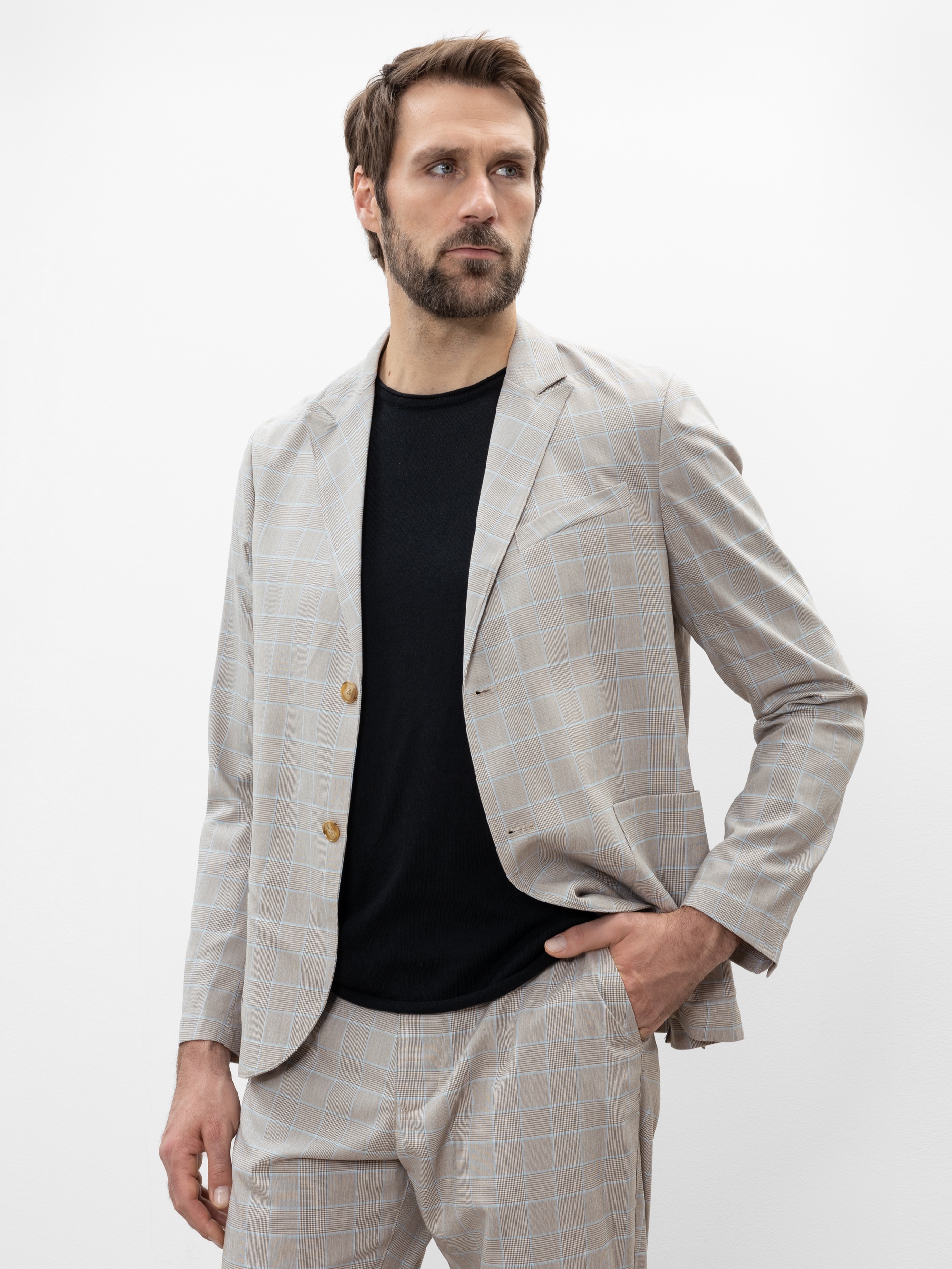 Ein Mann mit braunem Haar und Bart trägt einen hellgrau karierten Anzug mit einem schwarzen Hemd darunter. Er steht mit einer Hand in der Tasche und blickt leicht zur Seite vor einem schlichten weißen Hintergrund.
