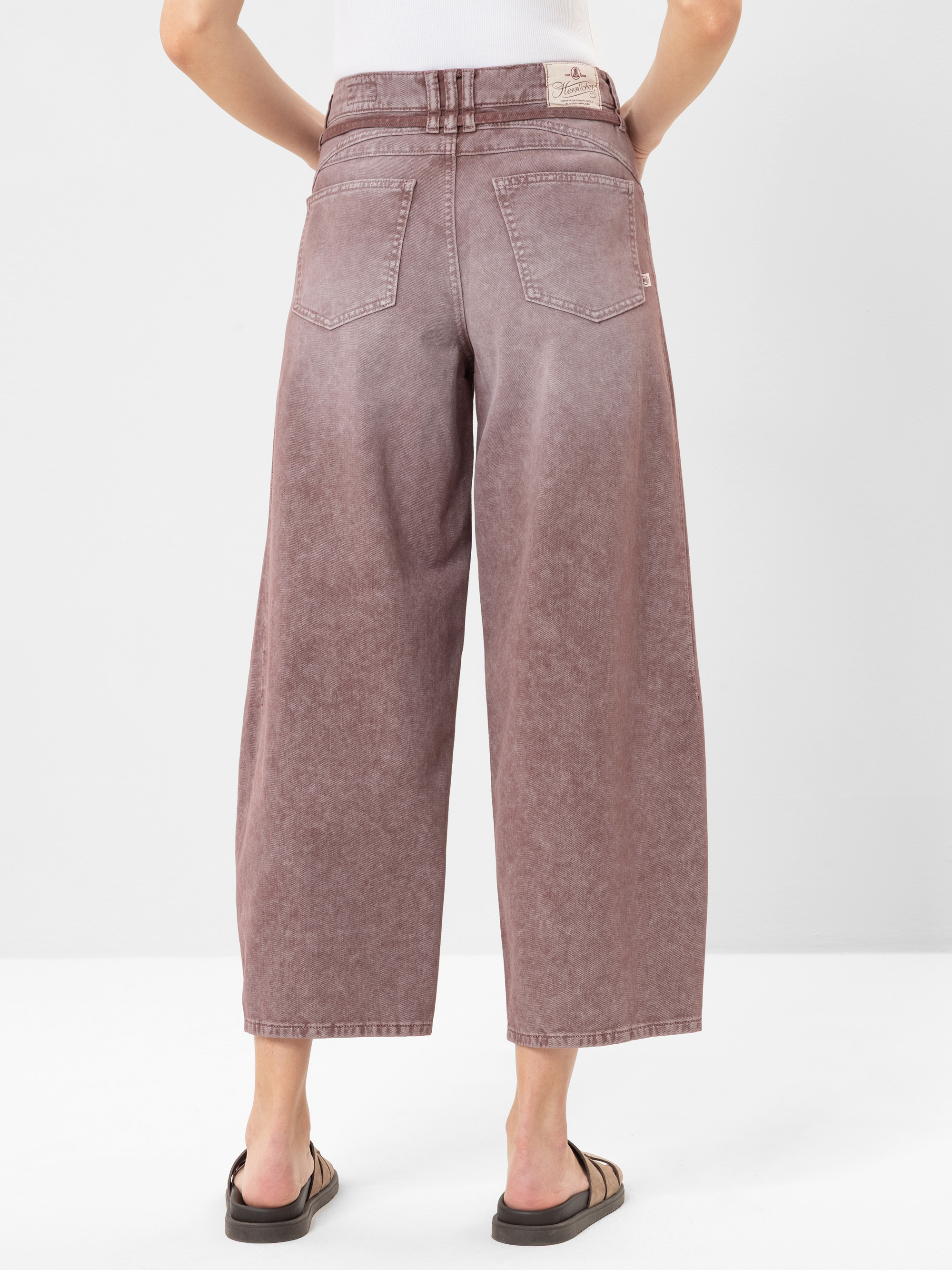 Eine Person trägt eine lilafarbene Jeans mit weitem Bein und verblasstem Finish, ein weißes Oberteil und schwarze Sandalen, von hinten vor einem schlichten weißen Hintergrund.