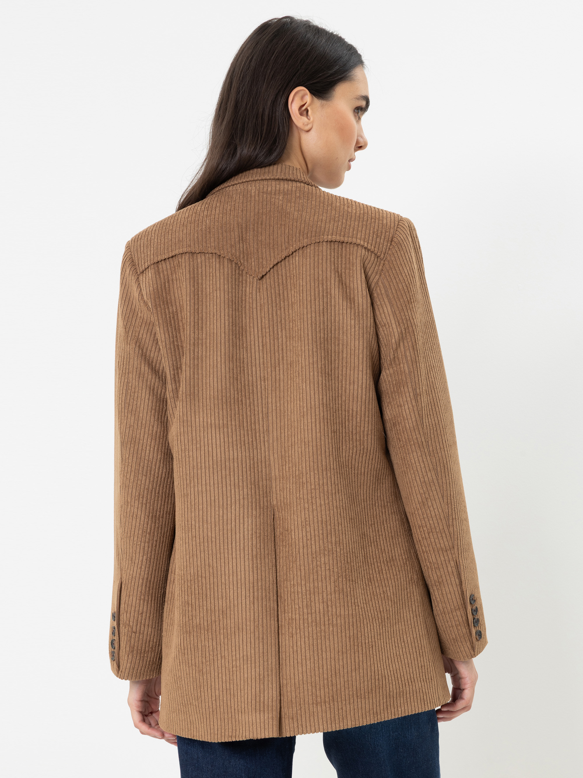 Eine Frau mit langen dunklen Haaren, die einen hellbraunen Cordblazer und eine dunkle Jeans trägt, ist von hinten vor einem schlichten weißen Hintergrund zu sehen.