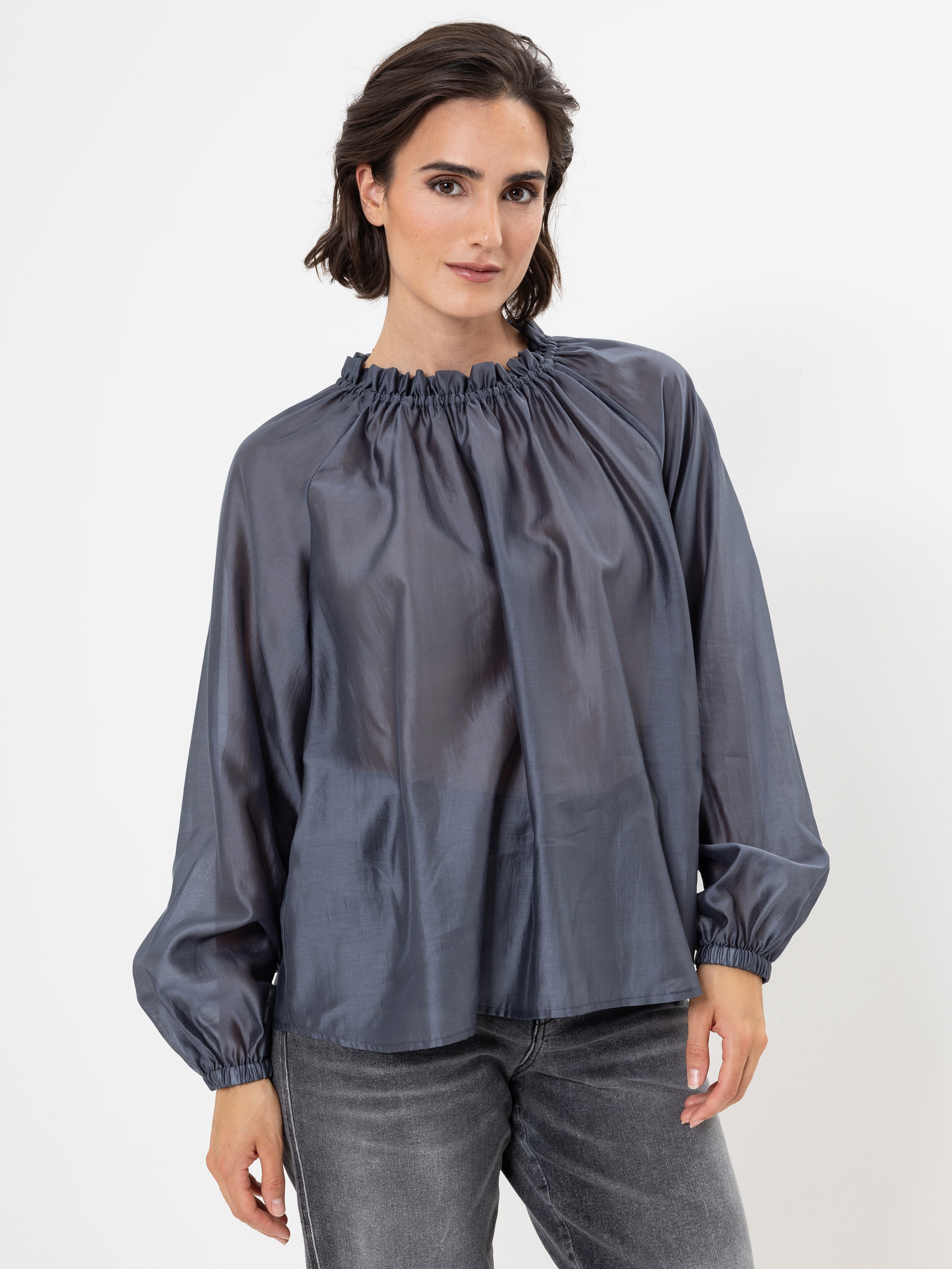 Eine Frau mit kurzen braunen Haaren, die eine locker sitzende, glänzende, langärmelige graue Bluse mit gerafftem Ausschnitt und graue Jeans trägt, steht vor einem schlichten weißen Hintergrund.