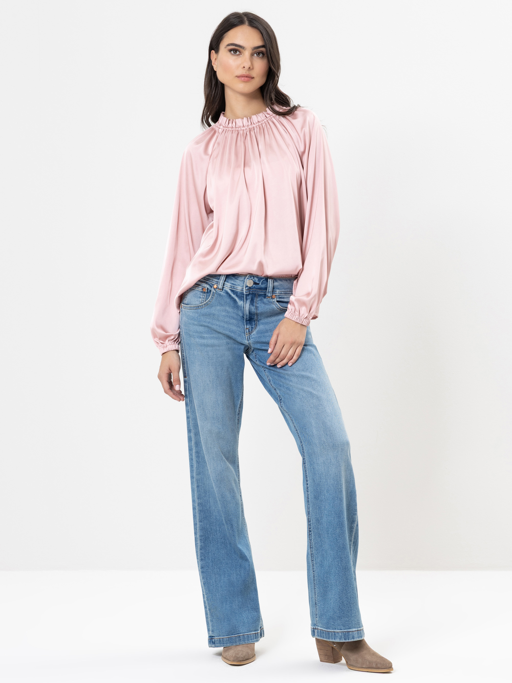 Eine Frau steht vor einem schlichten weißen Hintergrund und trägt eine hellrosa, langärmelige Bluse mit gerafftem Ausschnitt, blaue Jeans mit weitem Bein und beigefarbene Schuhe. Sie hat langes dunkles Haar und einen neutralen Gesichtsausdruck.