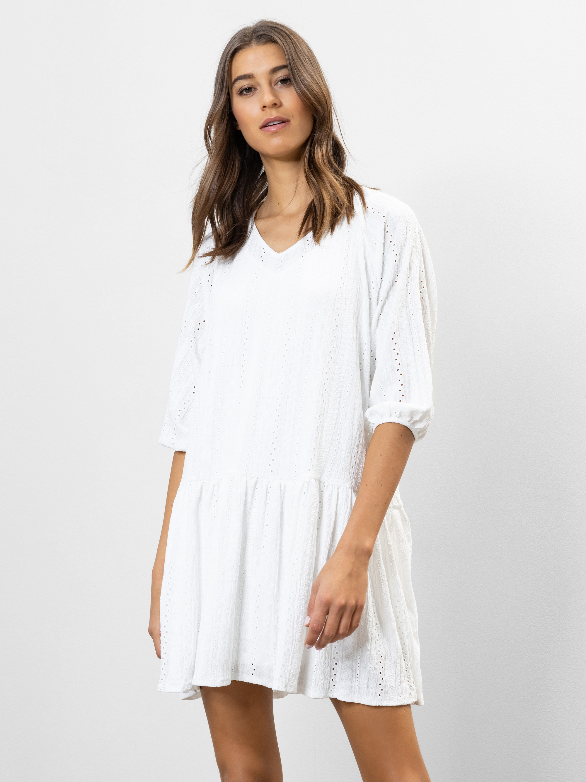 Eine Frau, die ein locker sitzendes, knielanges weißes Kleid mit V-Ausschnitt und Dreiviertelärmeln trägt, steht vor einem schlichten weißen Hintergrund.