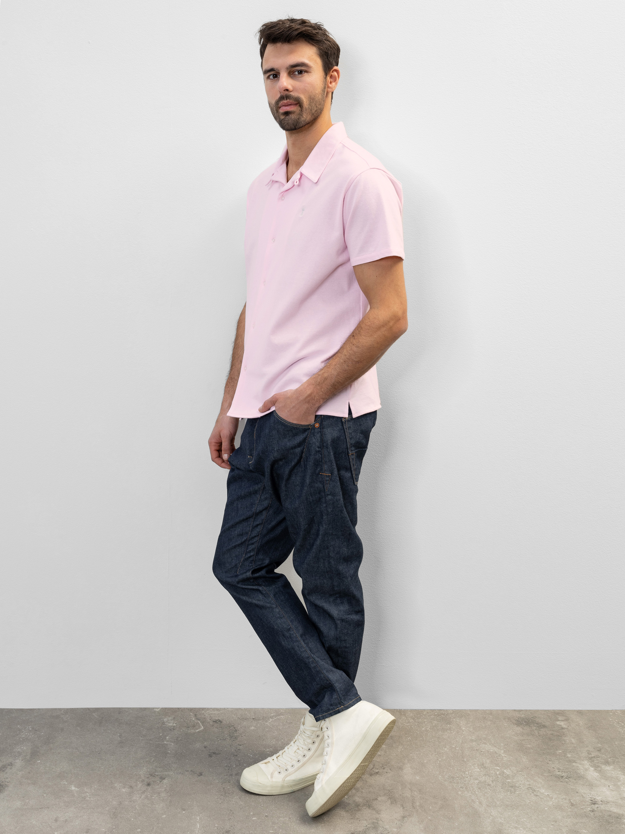 Ein Mann steht vor einer schlichten weißen Wand und trägt ein hellrosa, kurzärmeliges Polohemd, dunkelblaue Jeans und weiße Turnschuhe mit hohen Absätzen. Er schaut mit neutralem Blick in die Kamera.