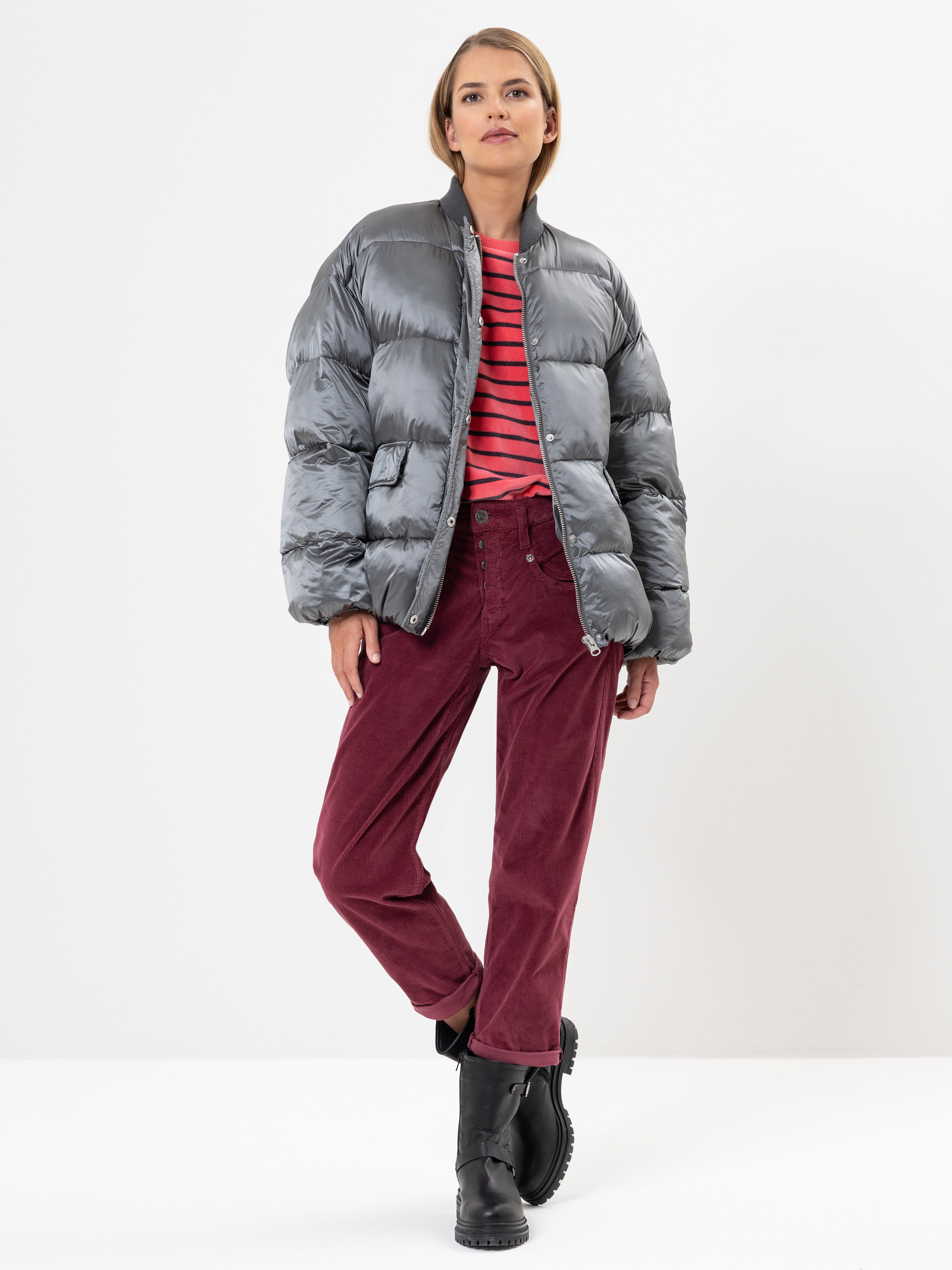 Eine Frau steht vor einem weißen Hintergrund und trägt eine glänzende graue Pufferjacke, ein rot-schwarz gestreiftes Hemd, eine rote Kordhose und schwarze Stiefel. Sie hat kurzes blondes Haar und einen neutralen Gesichtsausdruck.