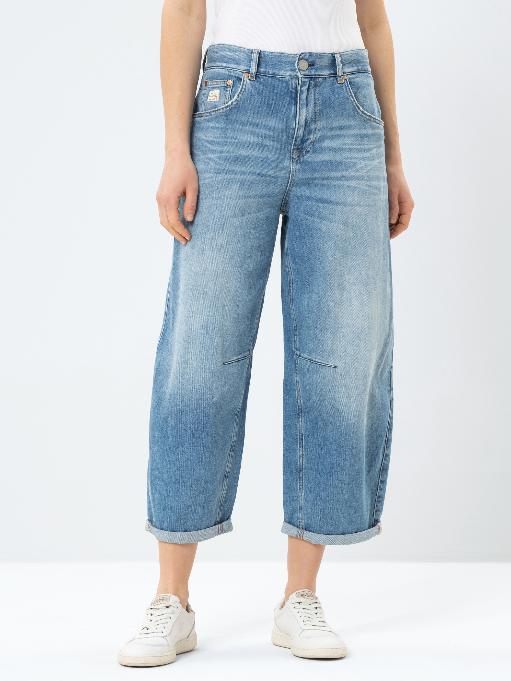 Eine Person, die eine hellblaue Jeans mit weitem Bein und Bündchen trägt, dazu ein weißes Oberteil und weiße Turnschuhe, steht vor einem schlichten hellen Hintergrund.