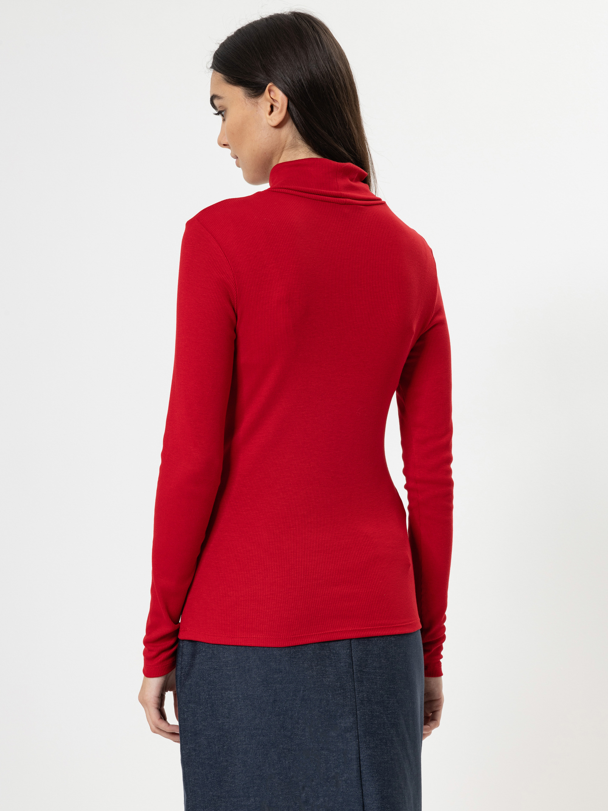 Eine Frau mit langen dunklen Haaren, von hinten gesehen, trägt einen roten Rollkragenpullover und einen dunkelblauen Rock vor einem schlichten weißen Hintergrund.