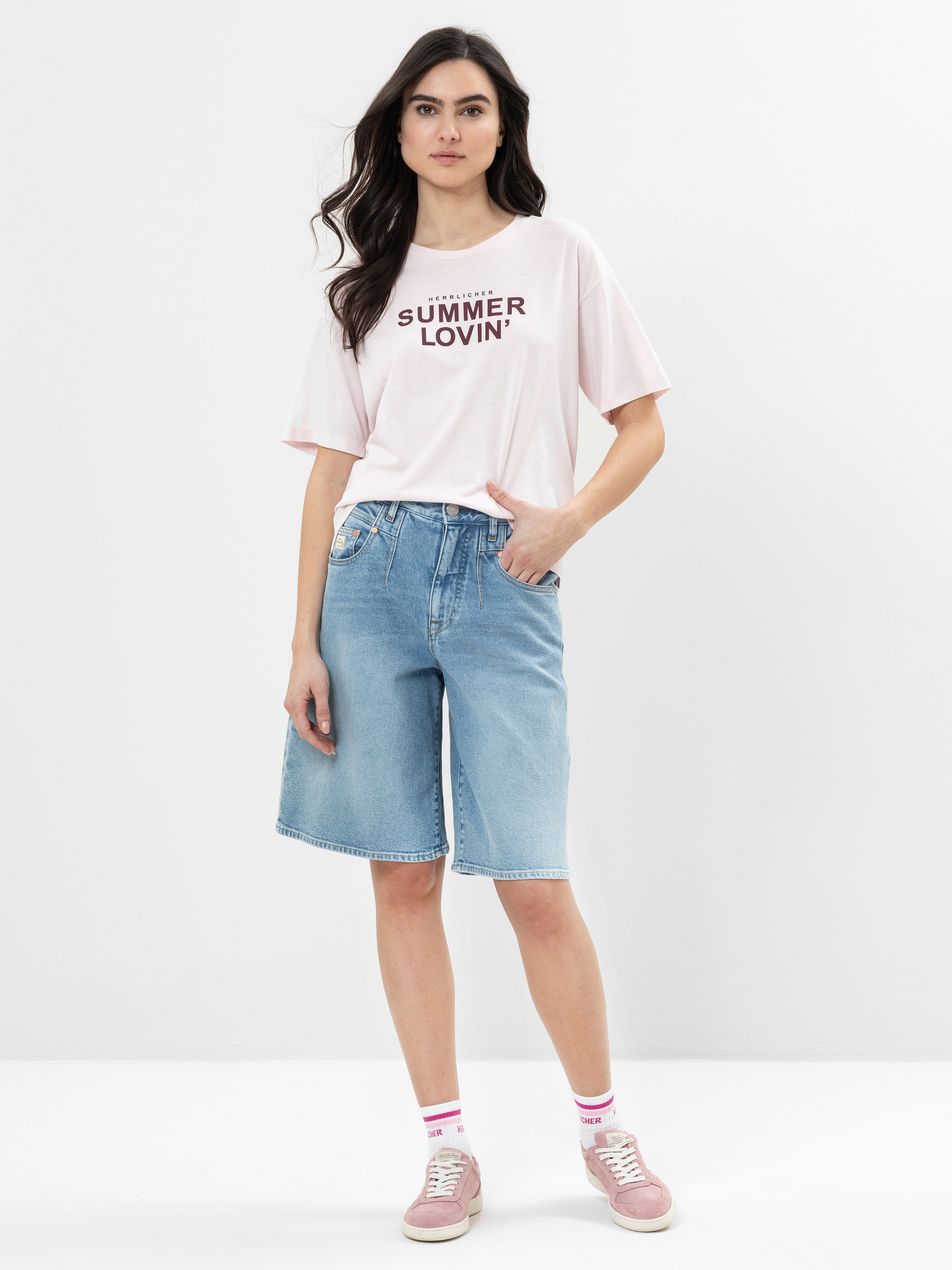 Eine Frau steht vor einem weißen Hintergrund und trägt ein hellrosa Summer Lovin-T-Shirt, knielange Jeans-Shorts, rosa Socken und rosa Turnschuhe. Sie hat langes dunkles Haar und einen neutralen Gesichtsausdruck.