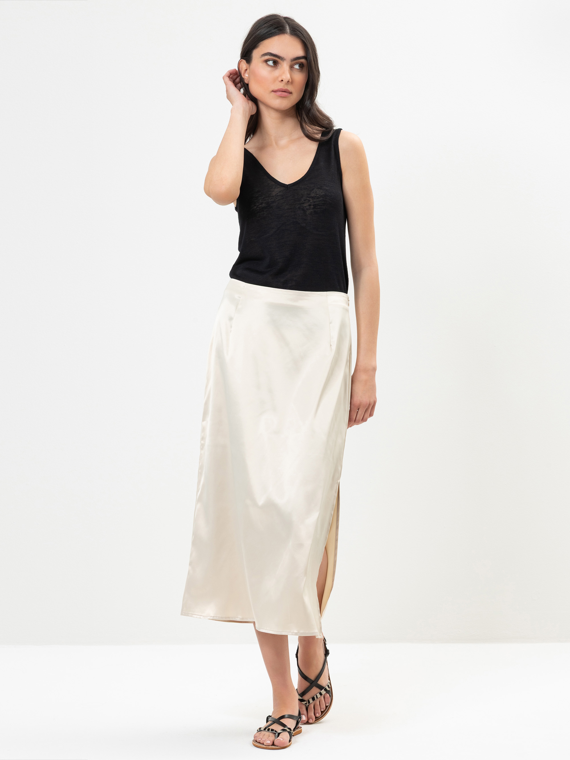 Eine Frau steht vor einem schlichten weißen Hintergrund und trägt ein ärmelloses schwarzes Oberteil, einen cremefarbenen seidigen Midirock mit Seitenschlitz und schwarze Riemchensandalen. Sie hat langes dunkles Haar und einen neutralen Gesichtsausdruck