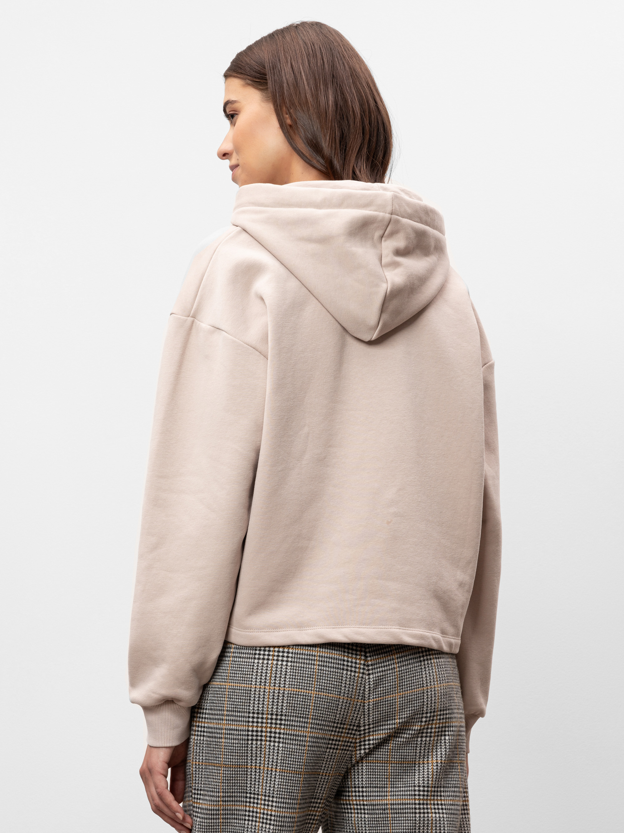 Eine Frau mit glattem braunem Haar trägt einen beigen Kapuzenpullover und eine karierte Hose und steht mit dem Rücken zur Kamera vor einem schlichten weißen Hintergrund.