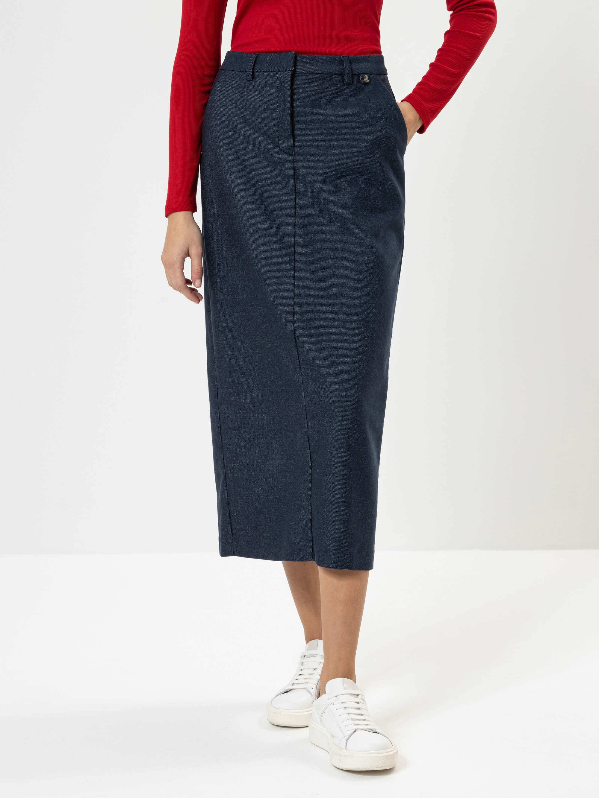 Eine Person, die ein rotes, langärmeliges Oberteil, einen hochgeschnittenen, dunkelblauen Midirock und weiße Turnschuhe trägt, steht vor einem schlichten weißen Hintergrund. Ihre Hände stecken in den Rocktaschen.