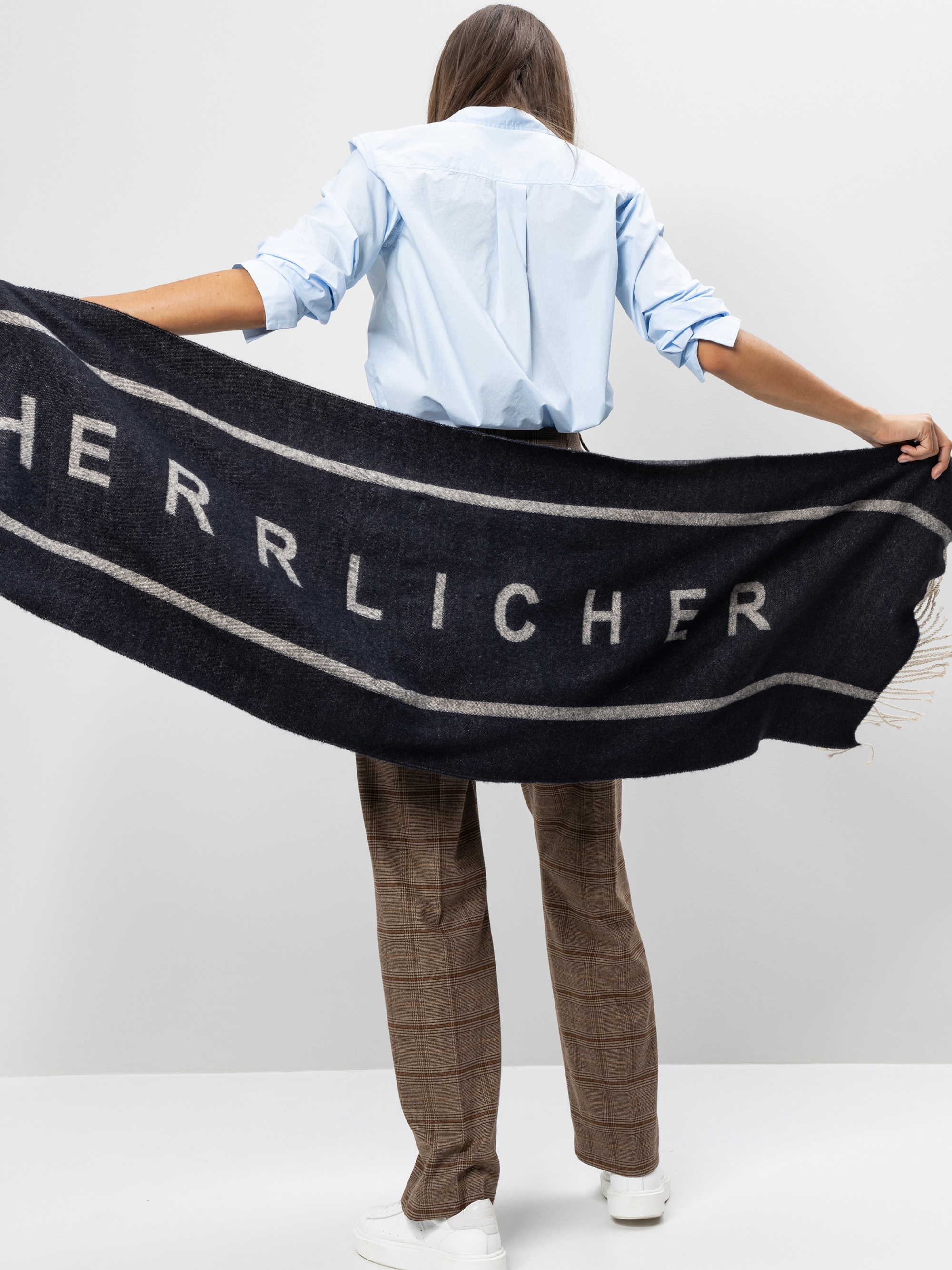 Eine Frau steht mit dem Gesicht zur Wand und hält einen großen dunklen Schal mit der Aufschrift HERRLICHER in der Hand. Sie trägt ein hellblaues Hemd, eine braun karierte Hose und weiße Turnschuhe vor einem schlichten Hintergrund.