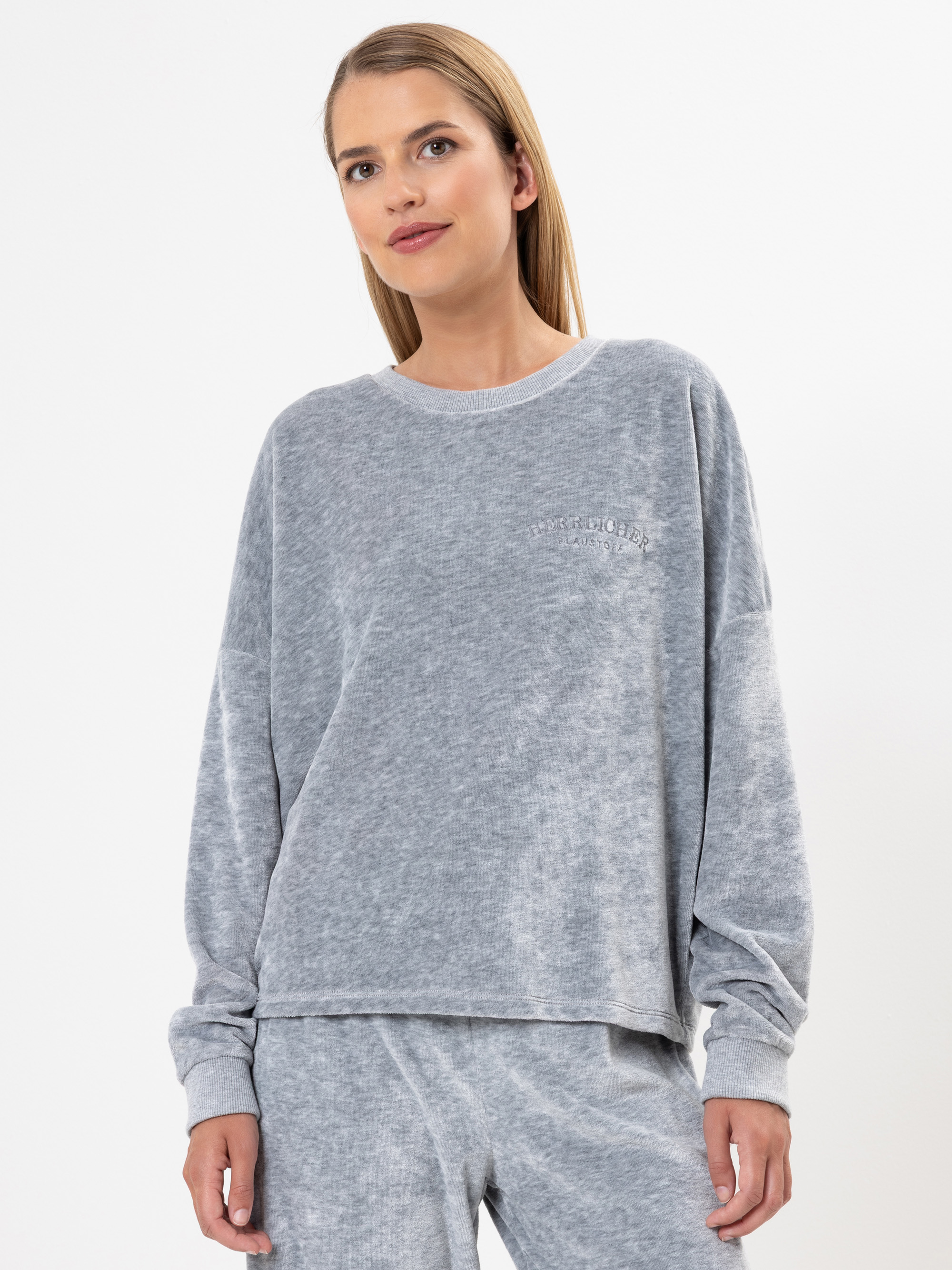 Eine Frau mit glattem, hellbraunem Haar trägt ein locker sitzendes, langärmeliges, hellgraues Sweatshirt und eine passende Hose. Sie steht vor einem schlichten weißen Hintergrund, blickt leicht zur Seite und lächelt sanft.