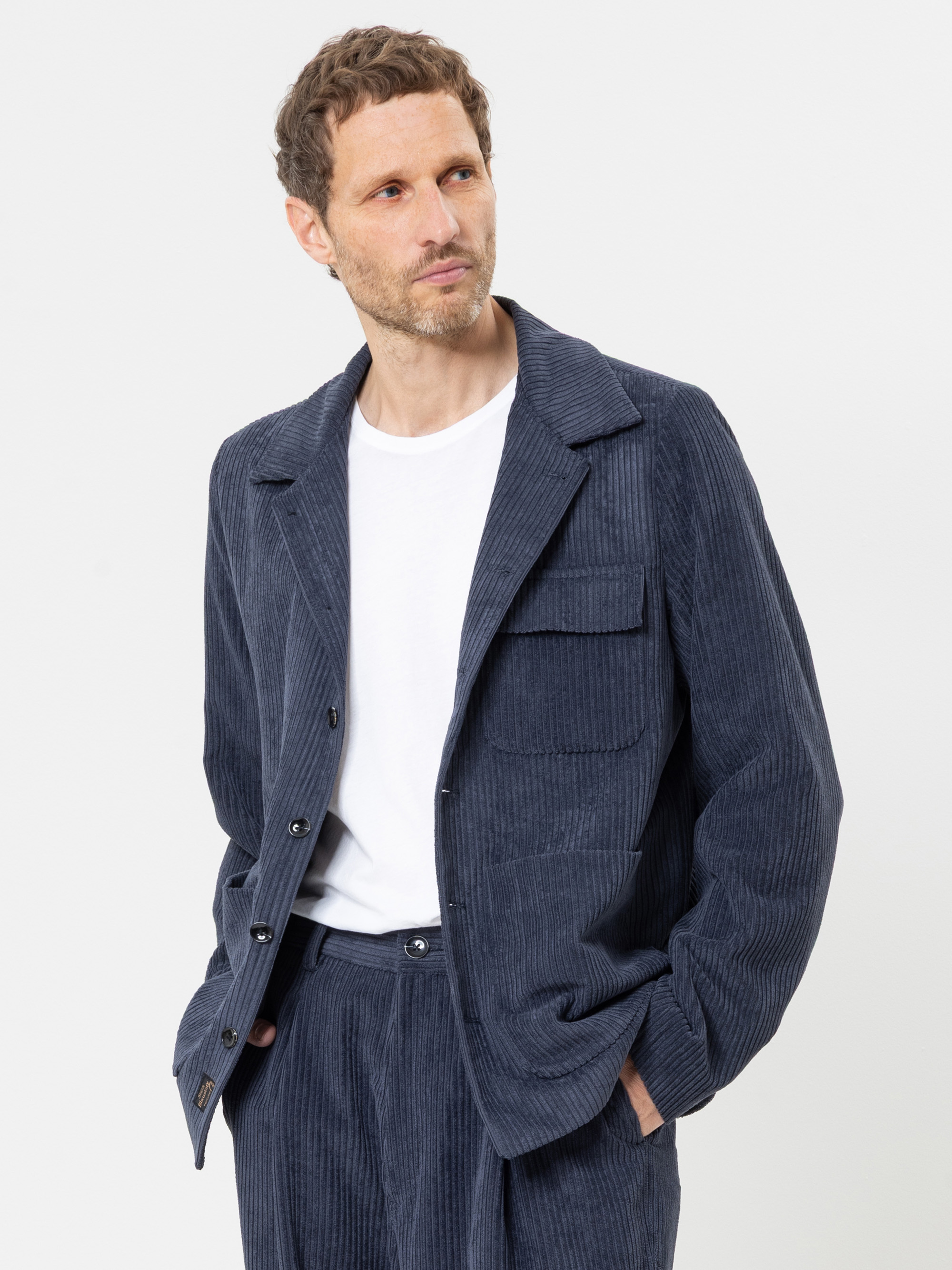 Ein Mann, der ein blaues Cord-Sakko und eine dazu passende Hose sowie ein weißes T-Shirt trägt, posiert mit den Händen in den Taschen vor einem schlichten weißen Hintergrund.