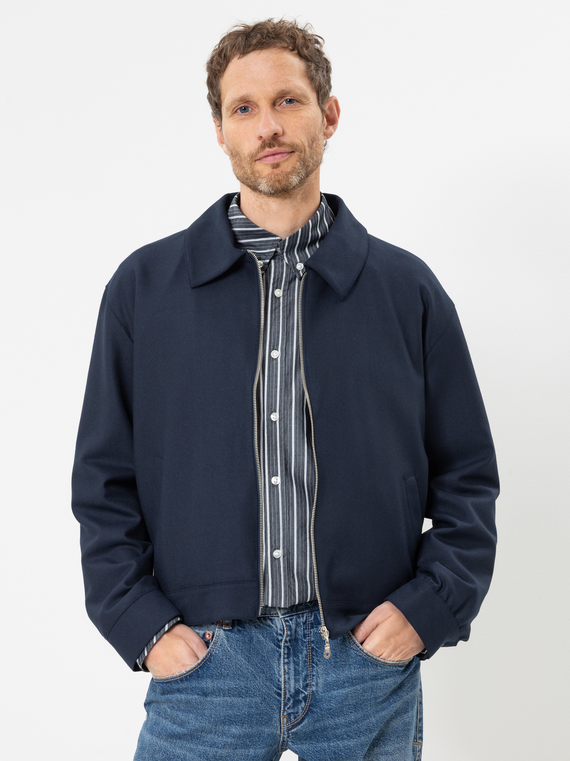 Ein Mann mit kurzen Locken und leichtem Bartwuchs trägt eine marineblaue Jacke über einem gestreiften Hemd mit Knöpfen sowie eine blaue Jeans; er steht vor einem schlichten weißen Hintergrund und blickt mit den Händen in den Taschen in die Kamera.