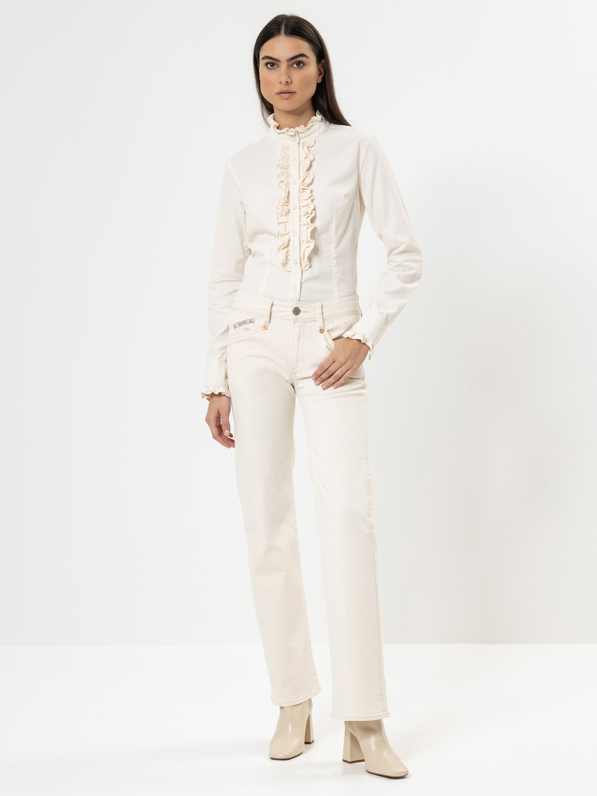 Eine Frau steht vor einem weißen Hintergrund und trägt ein weißes Outfit, bestehend aus einer gerüschten Bluse, einer hochtaillierten Hose und Stiefeletten. Sie hat langes dunkles Haar und einen neutralen Gesichtsausdruck, ihre linke Hand ruht an ihre