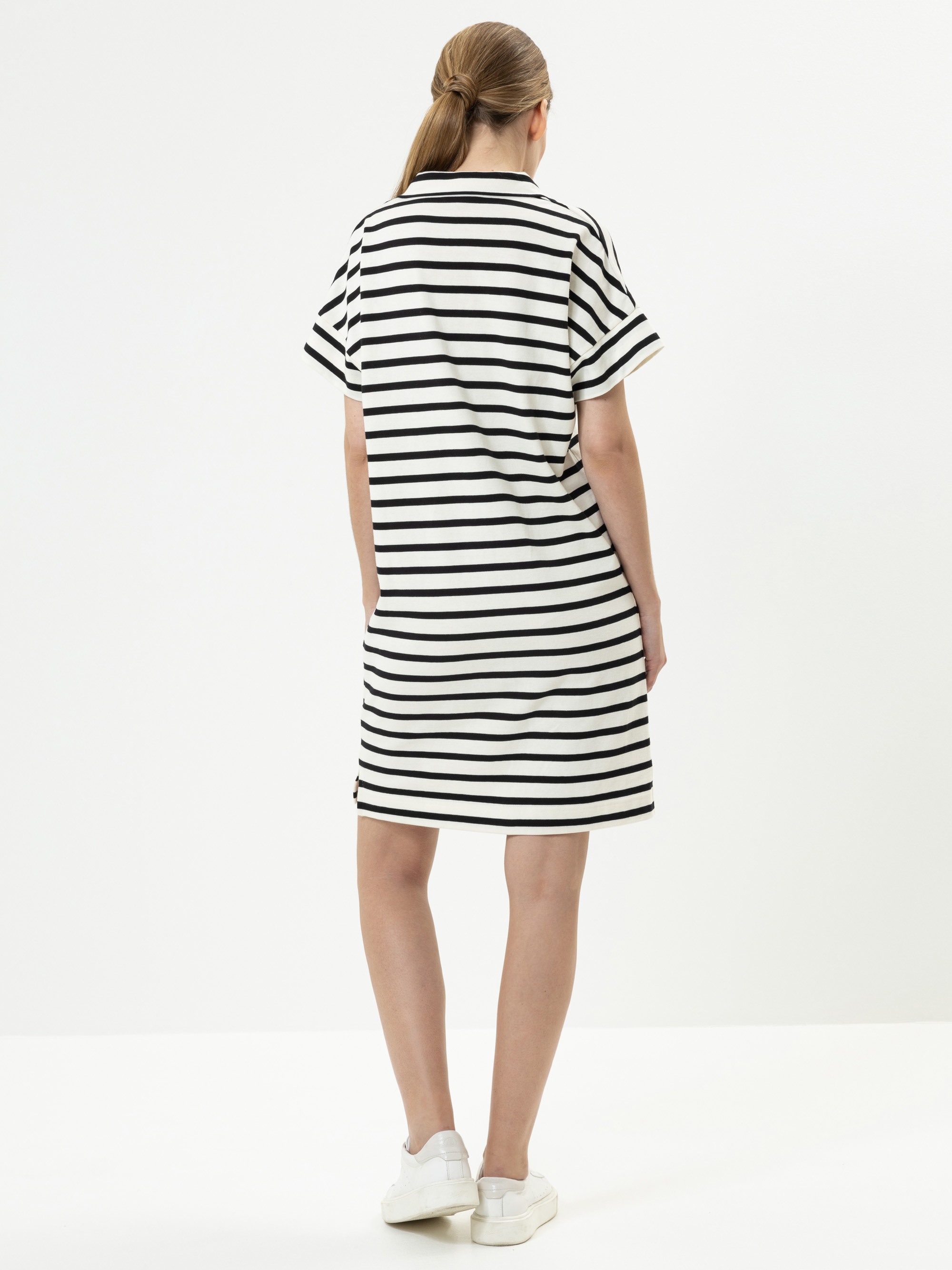 Eine Frau steht mit dem Gesicht zur Wand und trägt ein locker sitzendes, kurzärmeliges, knielanges Kleid mit schwarzen und weißen Querstreifen sowie weiße Turnschuhe. Ihr Haar ist zu einem tiefen Pferdeschwanz zurückgebunden. Der Hintergrund ist einf