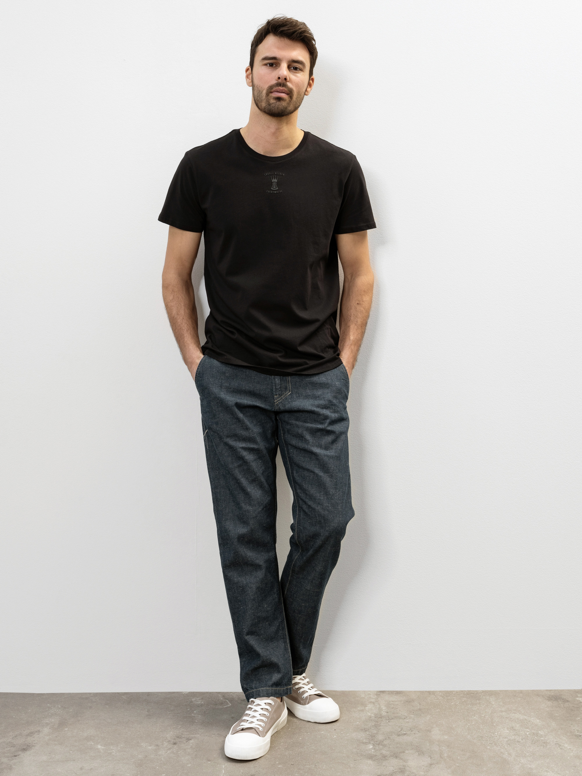 Ein Mann steht vor einer schlichten weißen Wand, trägt ein schwarzes T-Shirt, blaue Jeans und weiße Turnschuhe. Er hat die Hände in den Hosentaschen und schaut mit entspanntem Blick in die Kamera.