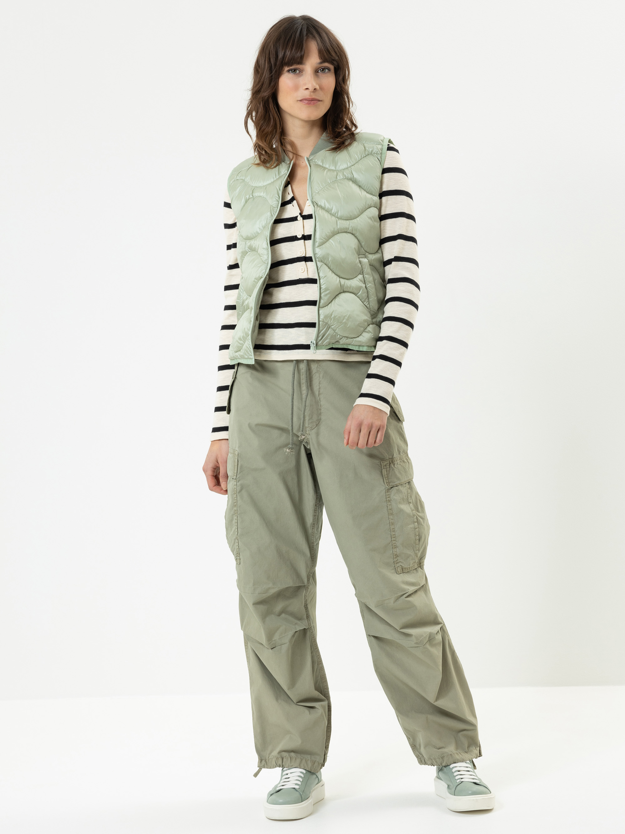 Eine Person steht vor einem schlichten weißen Hintergrund und trägt eine hellgrüne Steppweste, ein schwarz-weiß gestreiftes langärmeliges Hemd, eine weite olivfarbene Cargohose und hellgrüne Turnschuhe.