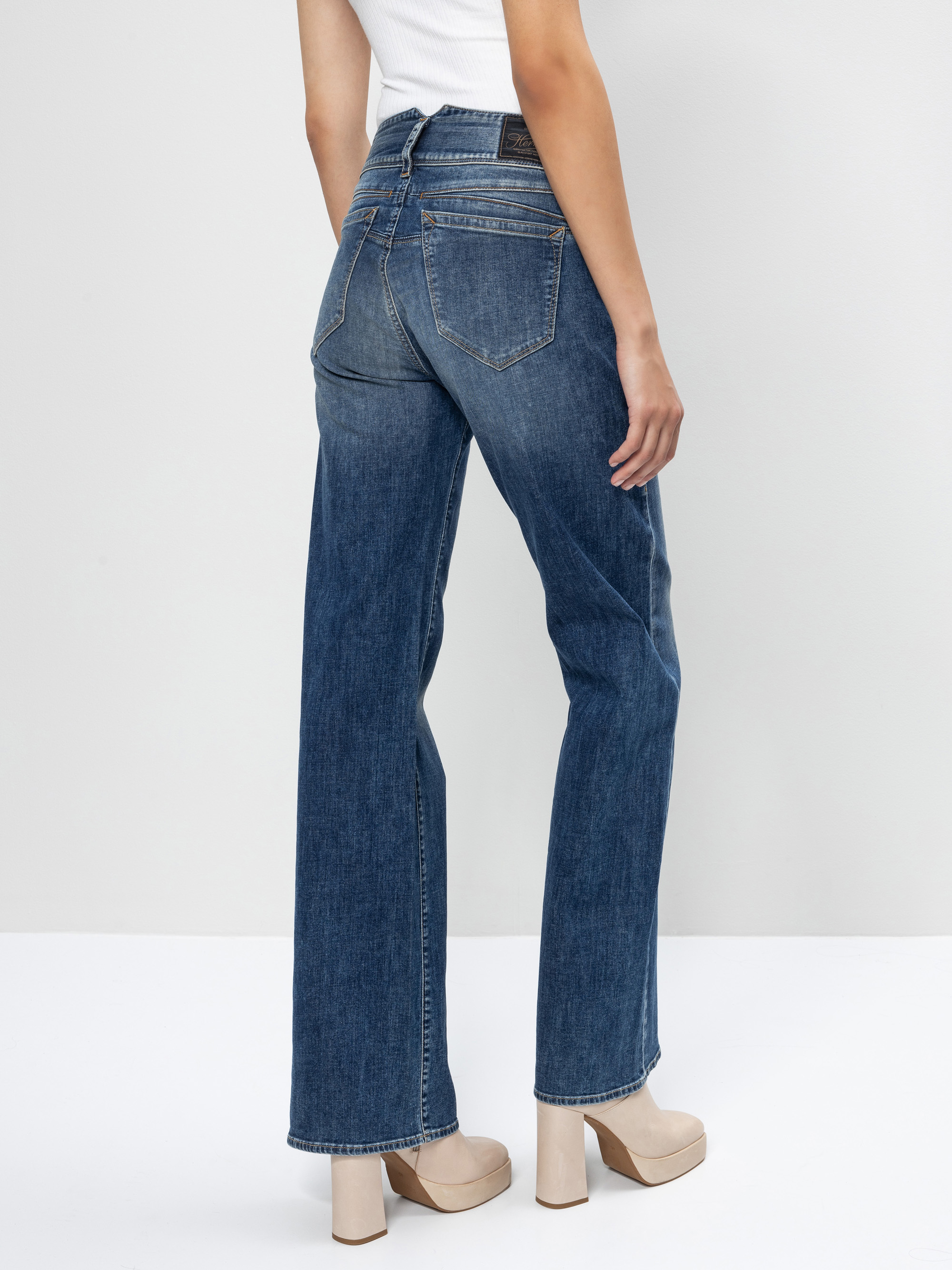 Eine Person steht mit dem Gesicht zur Wand, trägt eine hochgezogene blaue Jeans, ein weißes ärmelloses Oberteil und beigefarbene Plateauschuhe vor einem schlichten hellen Hintergrund.