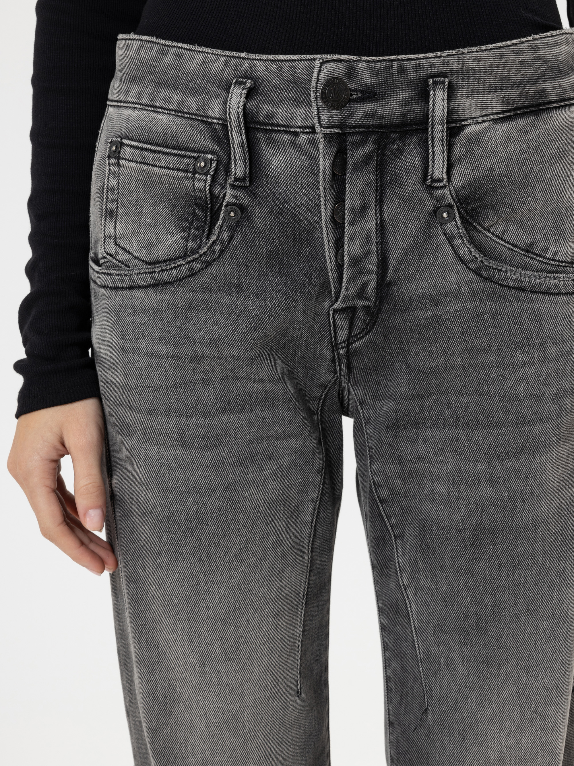 Eine Person, die eine hochgekrempelte, verblichene graue Jeans mit freiliegenden Knöpfen und ein schwarzes langärmeliges Rippenoberteil trägt, das von der Taille bis zur Mitte des Oberschenkels reicht.