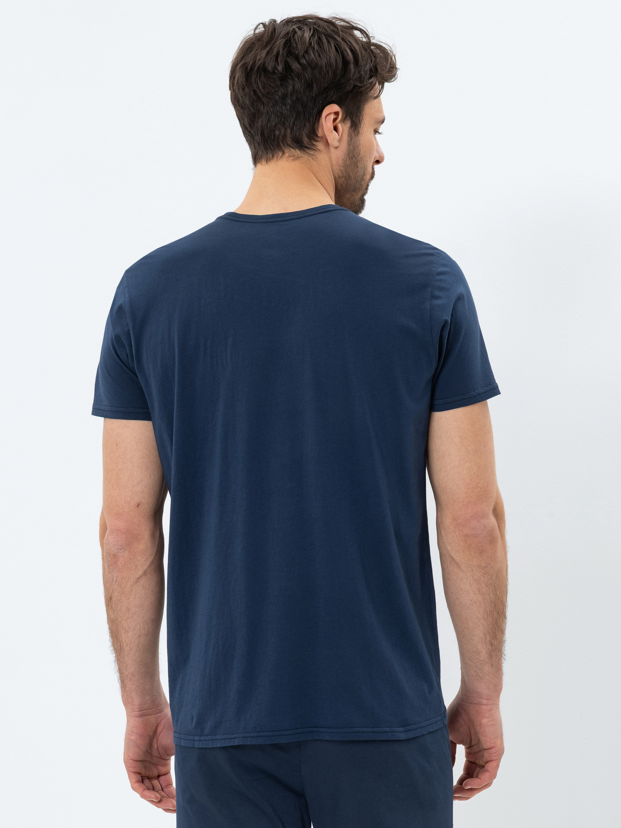 Ein Mann mit kurzen braunen Haaren ist von hinten zu sehen. Er trägt ein einfarbiges marineblaues T-Shirt und eine passende marineblaue Hose und steht vor einem weißen Hintergrund.