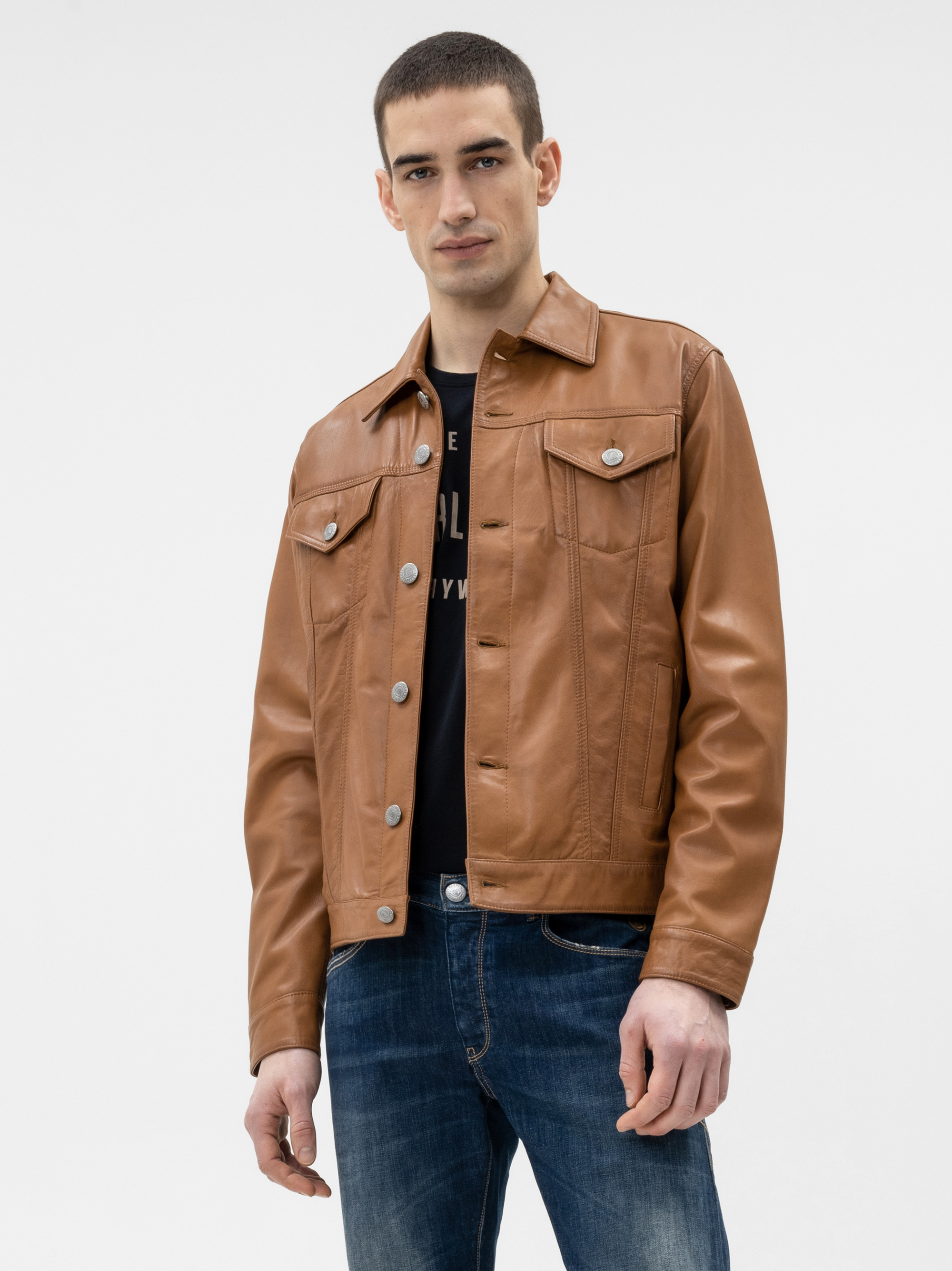 Ein Mann, der eine hellbraune Lederjacke über einem schwarzen Hemd und eine blaue Jeans trägt, steht vor einem schlichten weißen Hintergrund und schaut mit neutralem Blick direkt in die Kamera.