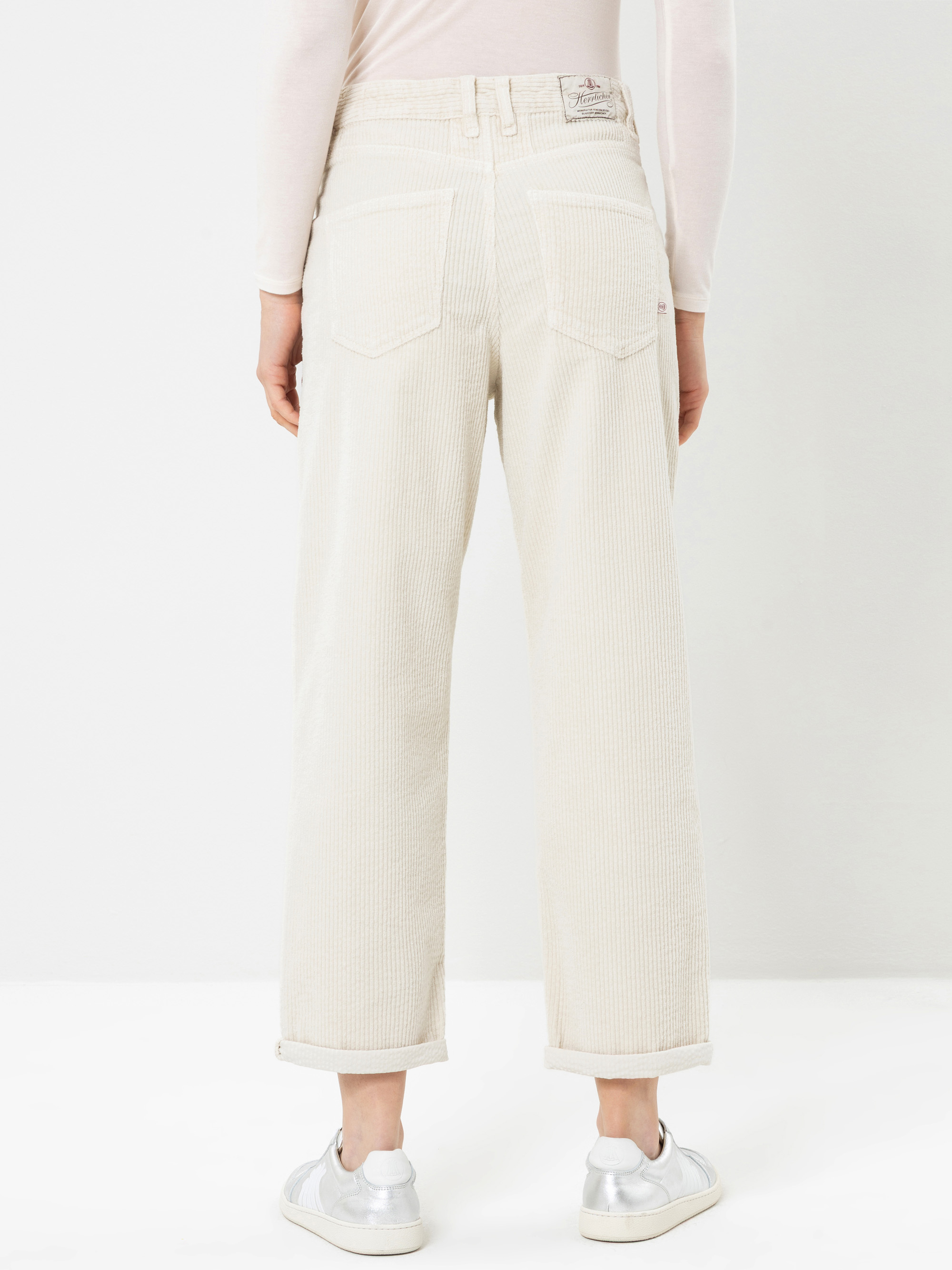 Eine Person, die mit dem Rücken zur Kamera steht und eine hellbeige Cordhose, ein helles Langarm-Oberteil sowie weiße Turnschuhe trägt. Die Hose hat umgeschlagene Säume und sichtbare Gesäßtaschen. Der Hintergrund ist einfarbig weiß.