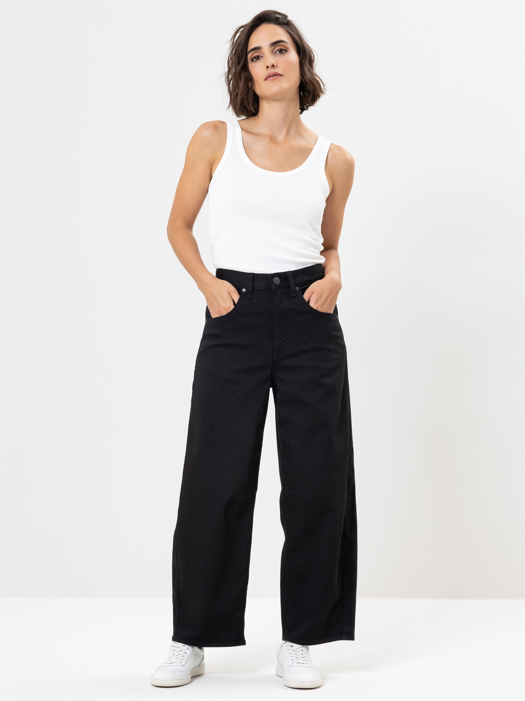 Eine Frau steht vor einem schlichten weißen Hintergrund, trägt ein weißes Tanktop, eine schwarze Hose mit weitem Bein und weiße Turnschuhe, die Hände in den Taschen und einen entspannten Gesichtsausdruck.