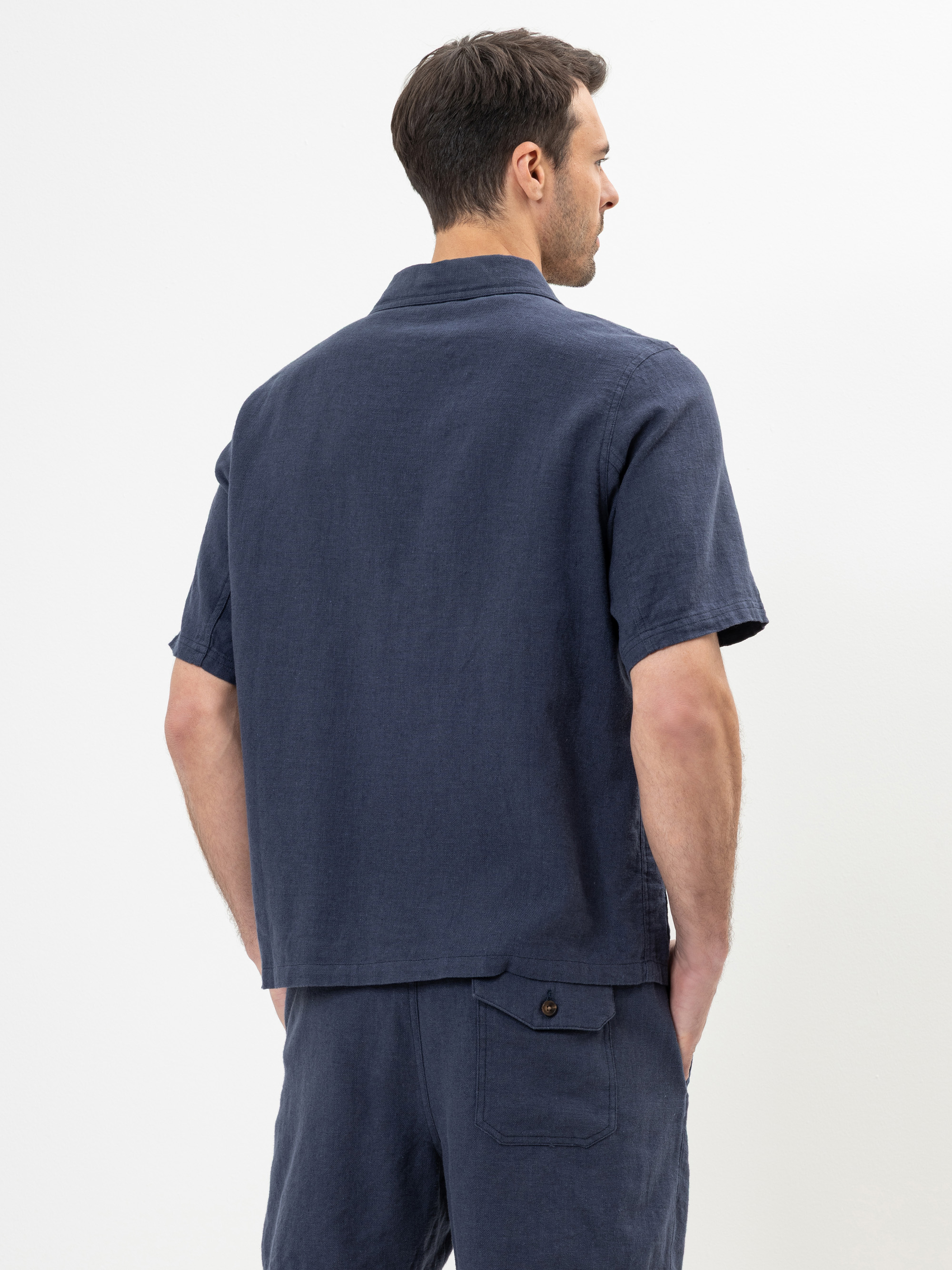 Ein Mann steht mit dem Rücken zur Kamera und trägt ein marineblaues kurzärmeliges Hemd und passende Shorts. Seine Hände stecken in den Taschen, und er steht vor einem schlichten weißen Hintergrund.