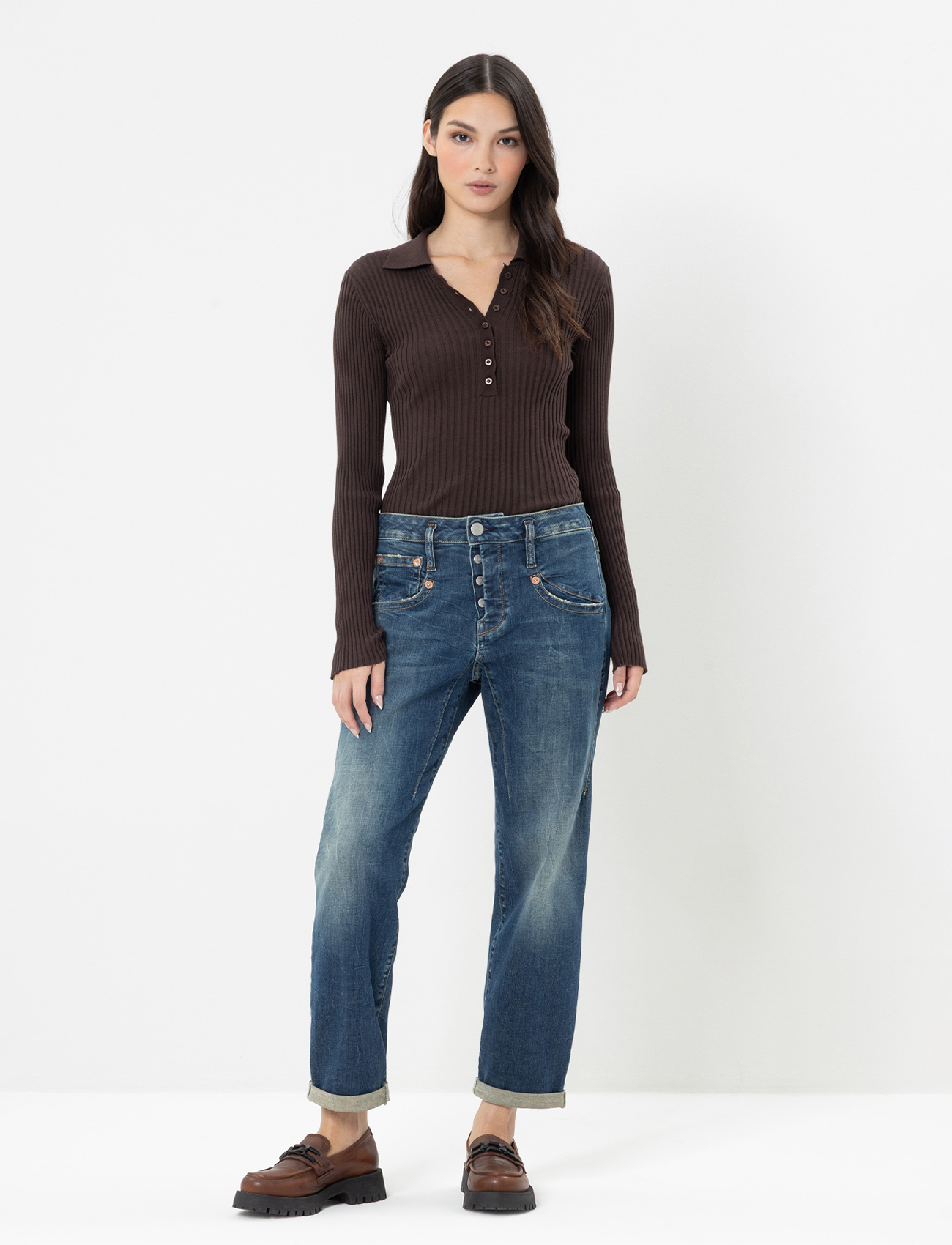 Eine Frau steht vor einem schlichten weißen Hintergrund und trägt eine abgeschnittene blaue Jeansjacke und passende blaue Jeans mit aufgerollten Bündchen. Sie hat langes dunkles Haar und ist barfuß, abgesehen von schwarzen Schleifen-Sandalen.