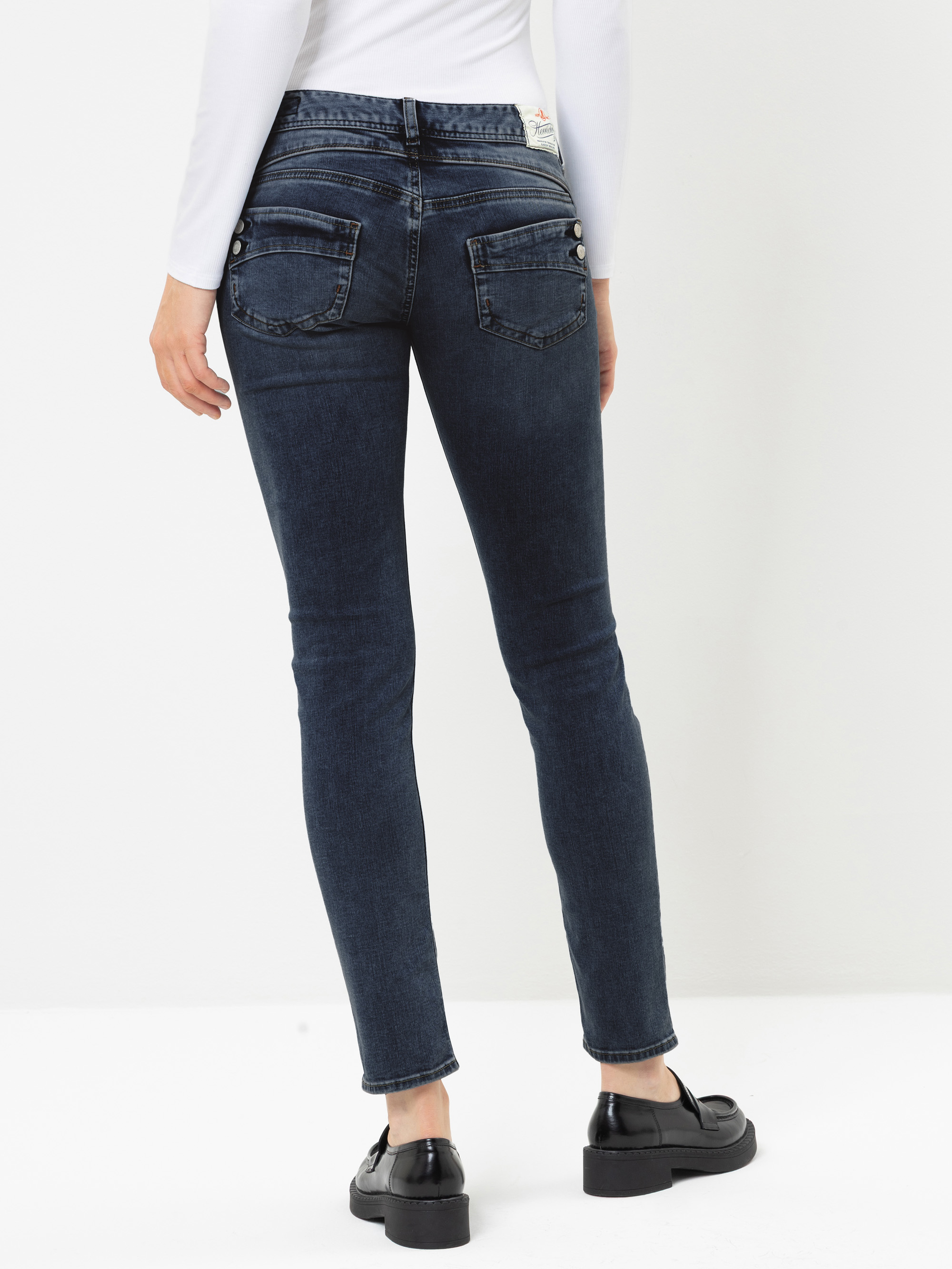 Eine Person, die eine dunkelblaue Skinny-Jeans, schwarze Slipper und ein weißes Langarm-Oberteil trägt, von hinten gesehen, vor einem einfarbigen weißen Hintergrund stehend.