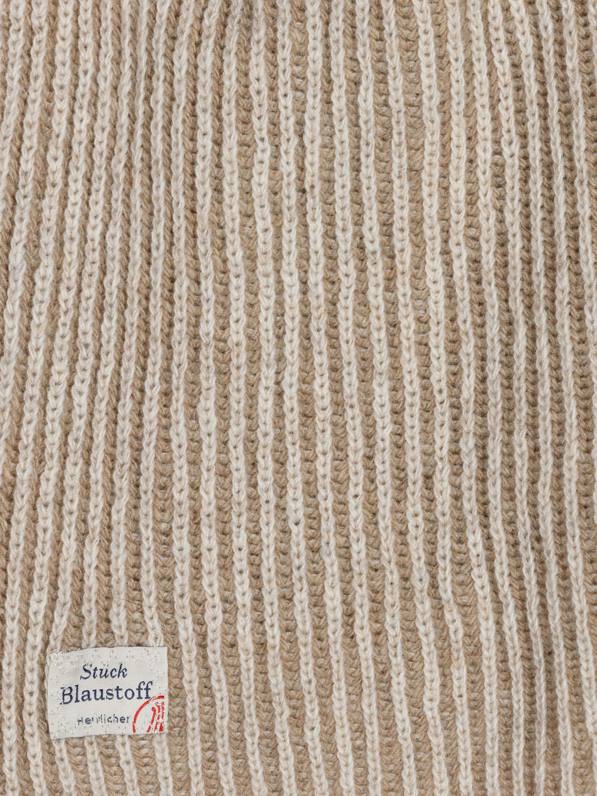 Nahaufnahme eines Gewirkes mit abwechselnd beigen und weißen Längsstreifen. In den Stoff ist ein kleines weißes Etikett eingenäht, das die Aufschrift Stück Blaustoff Herrlicher und eine rote stempelartige Markierung trägt.