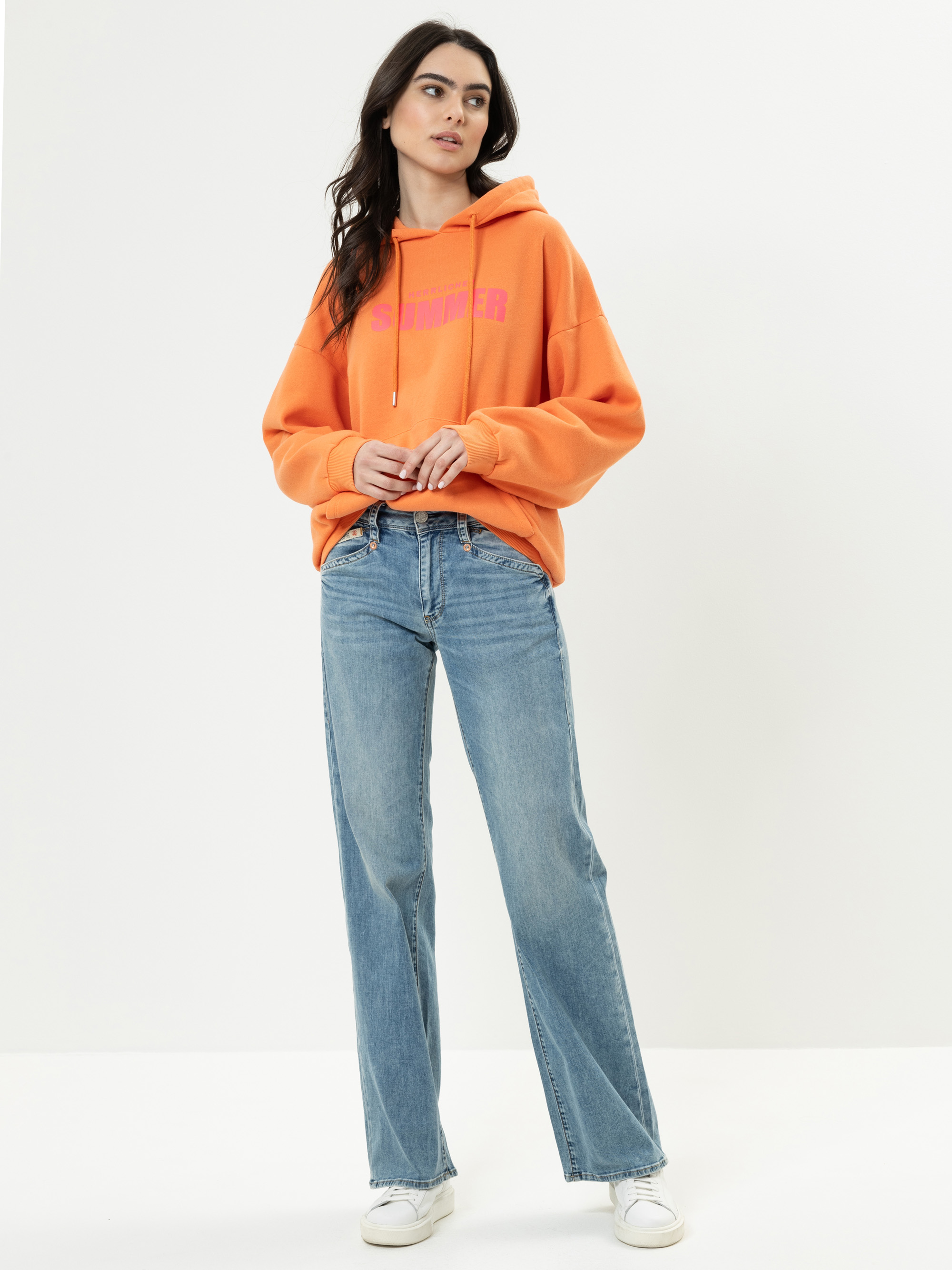 Eine Frau steht vor einem schlichten weißen Hintergrund und trägt einen orangefarbenen Kapuzenpullover mit Text, blaue Jeans mit geradem Bein und weiße Turnschuhe. Sie hat langes dunkles Haar und einen neutralen Gesichtsausdruck, ihre Hände ruhen an i