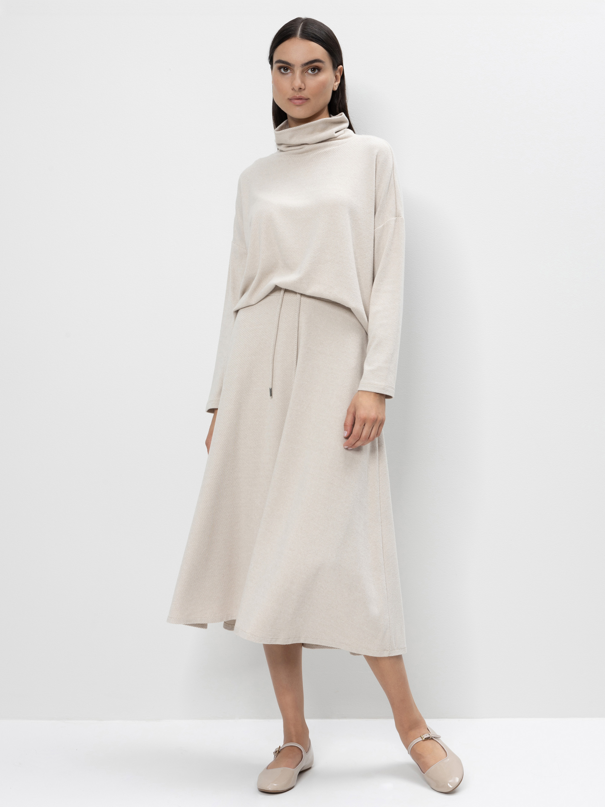 Eine Frau steht vor einem schlichten weißen Hintergrund und trägt einen lockeren, beigen Rollkragenpullover und einen passenden Midirock mit Bindeband in der Taille. Sie kombiniert das Outfit mit hellbeigen Flats und hat glattes, dunkles Haar.