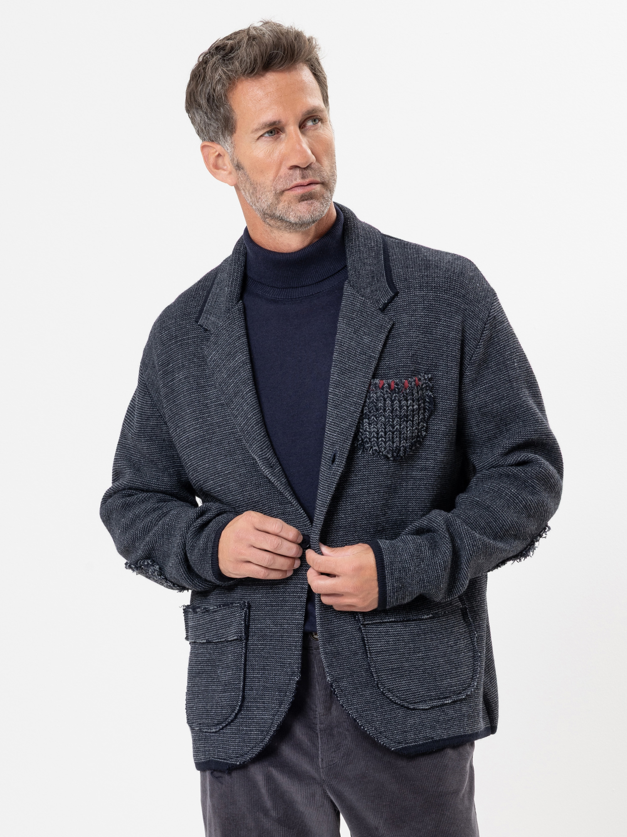 Ein Mann mit kurzem braunem Haar und gestutztem Bart trägt einen dunkelnavyfarbenen Rollkragenpullover und einen strukturierten dunkelgrauen Blazer mit aufgesetzten Taschen vor einem schlichten weißen Hintergrund.